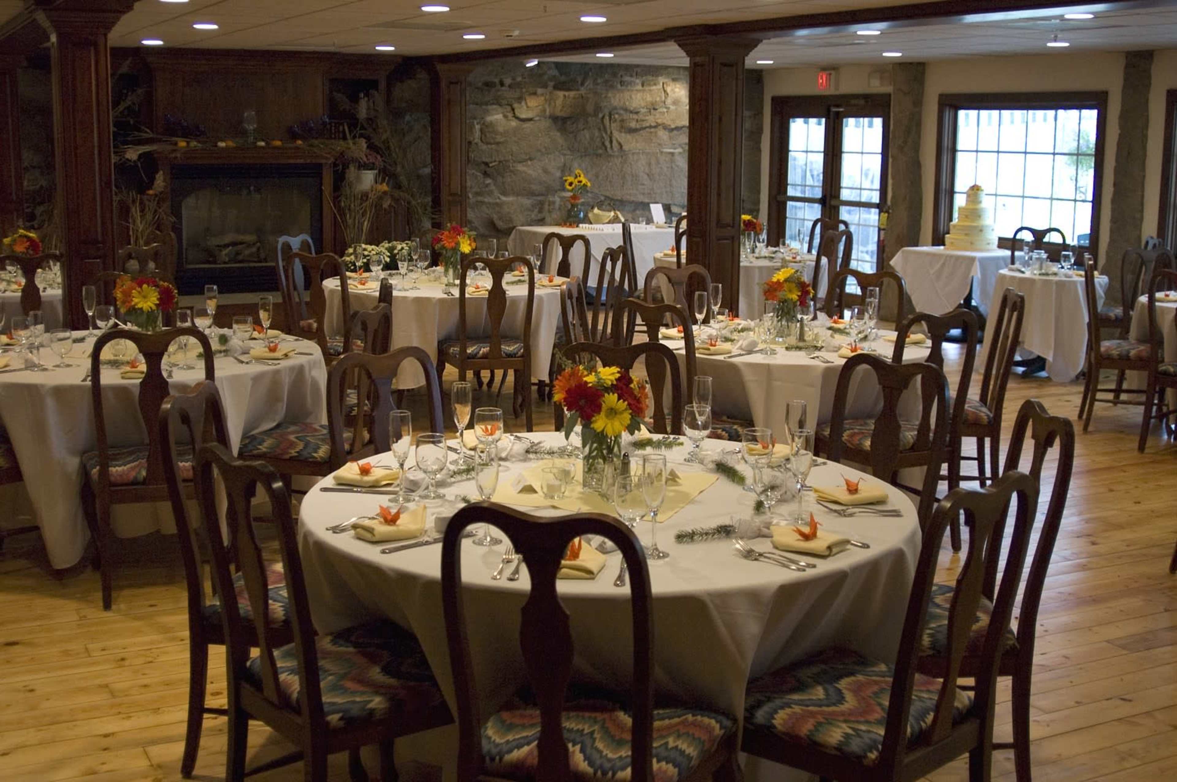 A dining room is set with several round tables adorned with white tablecloths and floral centerpieces, accompanied by chairs arranged around each table.