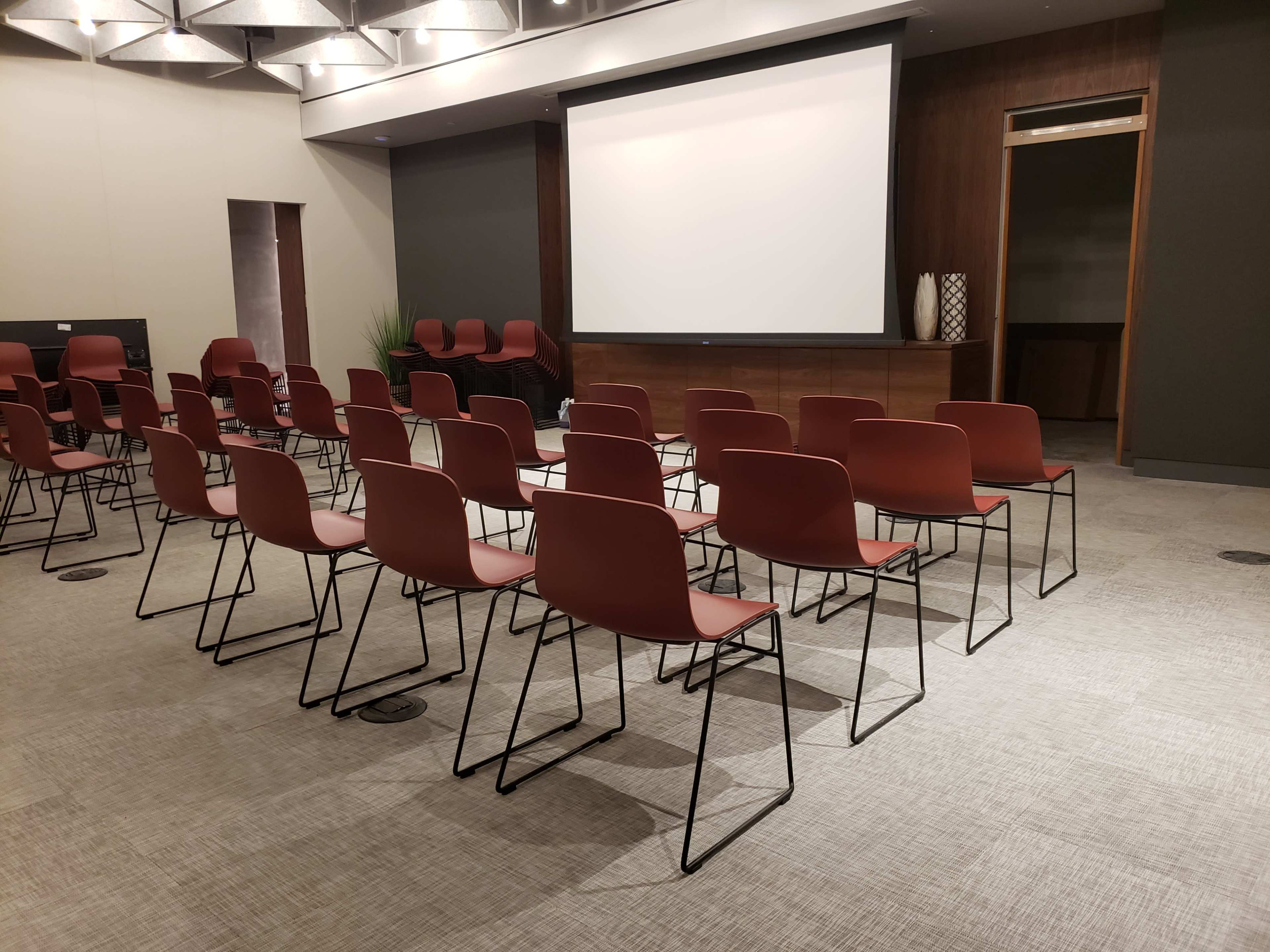 A row of empty red chairs is arranged in a meeting room facing a blank projection screen.