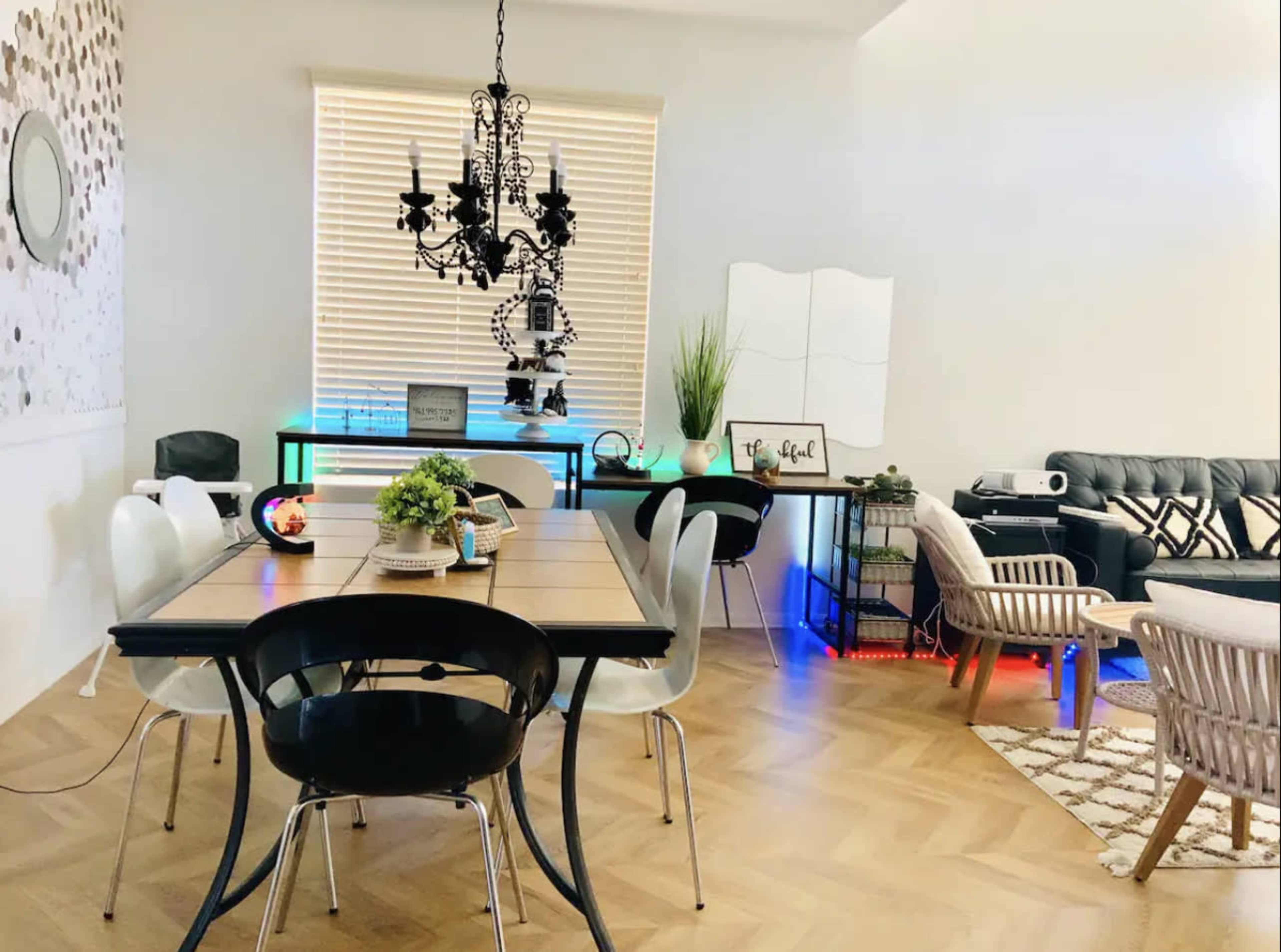 A dining area features a wooden table surrounded by black and white chairs, with a modern chandelier above and decorative plants on a side table.