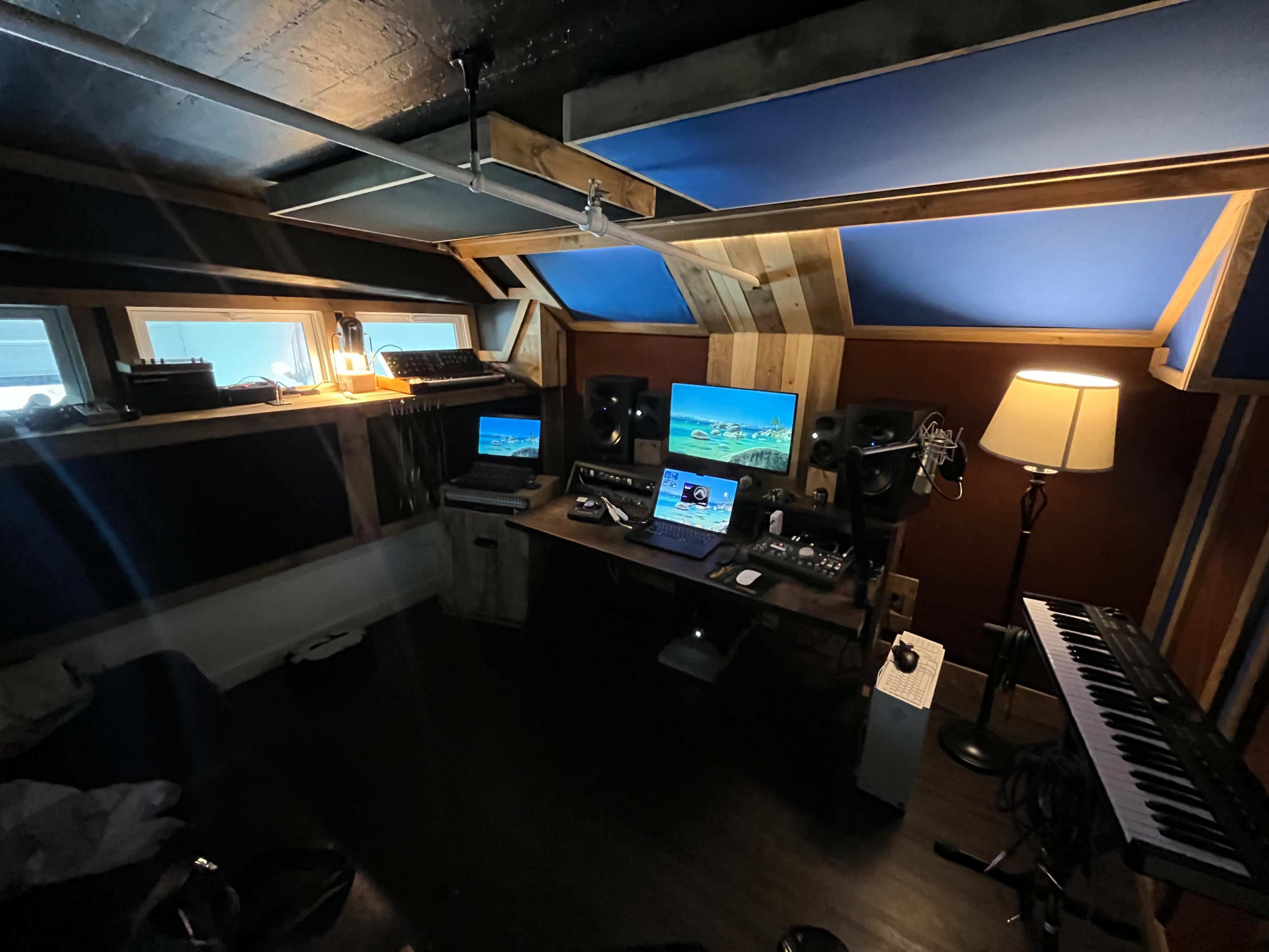 The image shows a recording studio with a central desk featuring two computer monitors, audio equipment, and a keyboard, surrounded by wooden paneling and soft lighting.