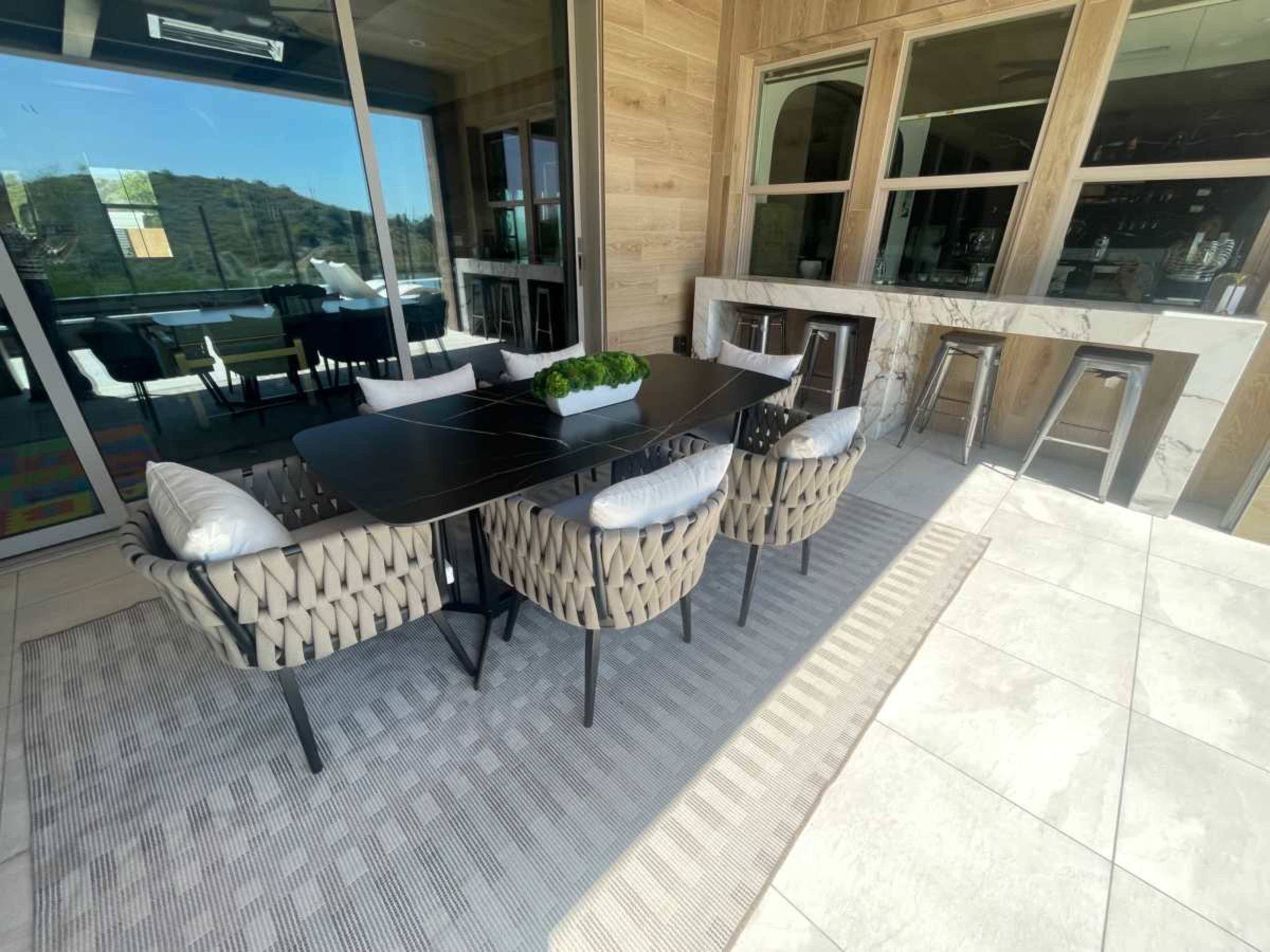 An outdoor dining area with a dark table surrounded by woven chairs and a bar area featuring marble countertops.
