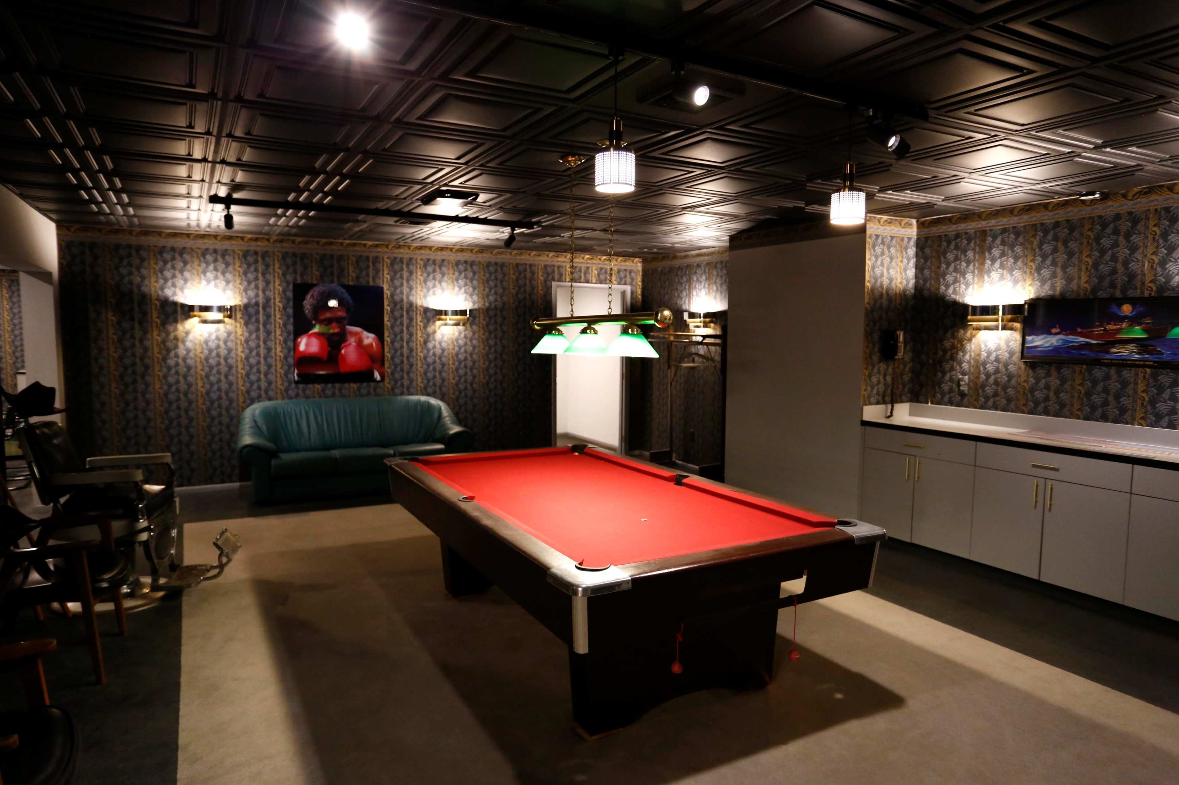 The room features a red billiard table at the center, surrounded by a green couch and a bar area, with patterned wallpaper and modern lighting.