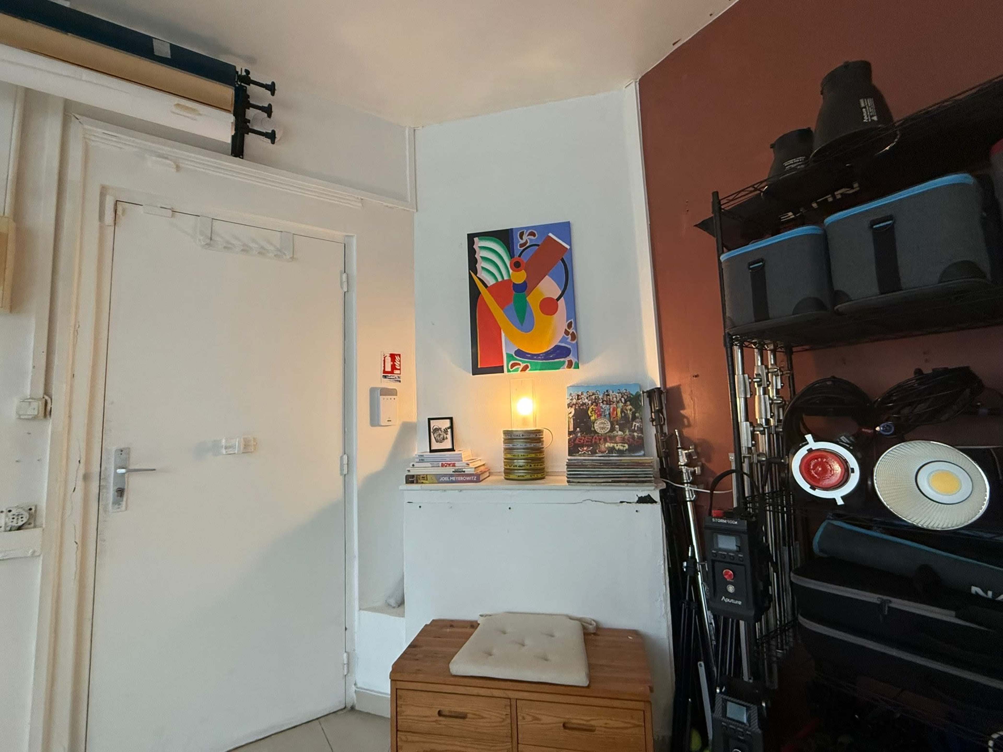 The image shows a corner of a room with a door, a colorful artwork on the wall, and a shelf stacked with books and equipment.