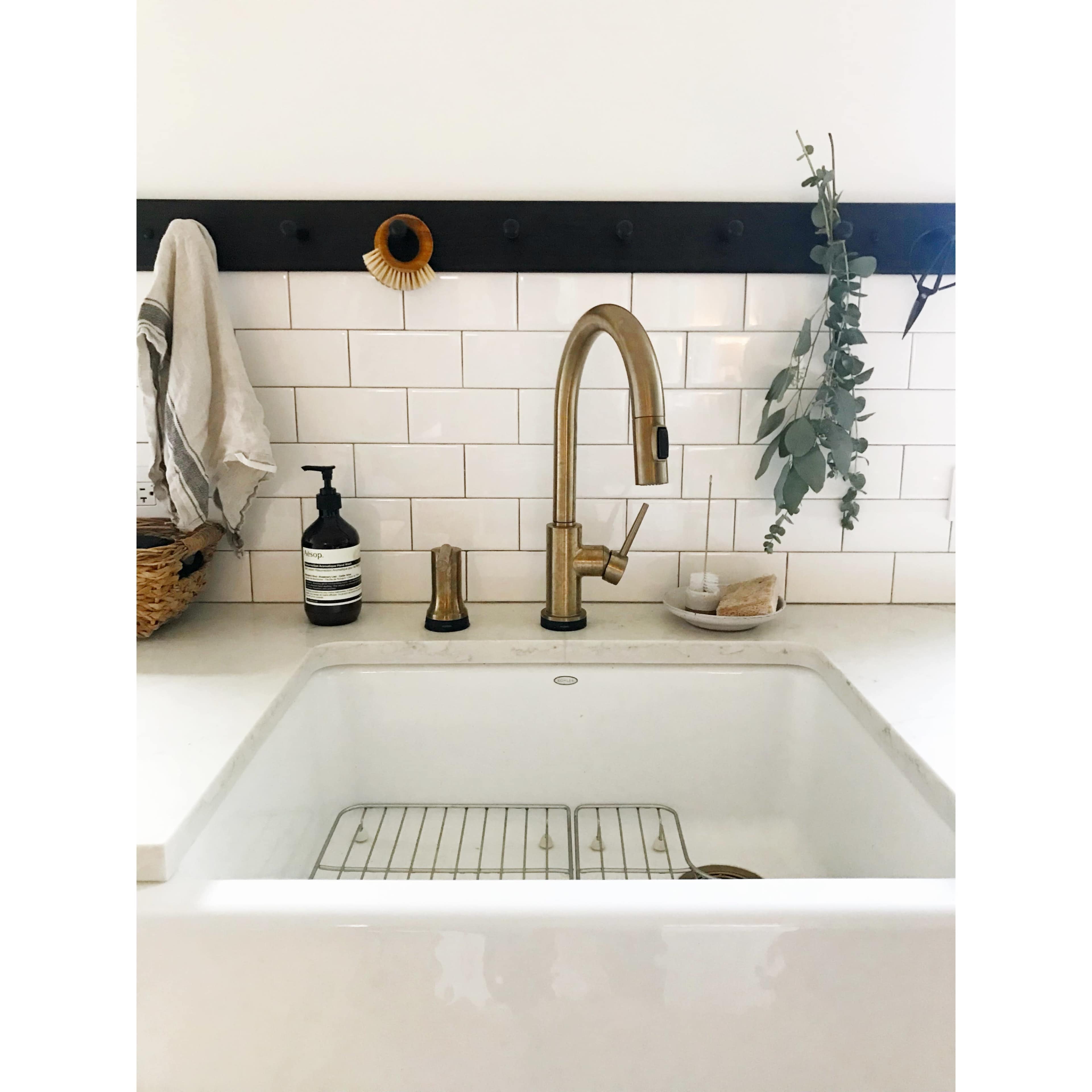 A modern kitchen sink features a brass faucet, white tiled backsplash, and neatly arranged accessories, including a soap dispenser and a hanging plant.