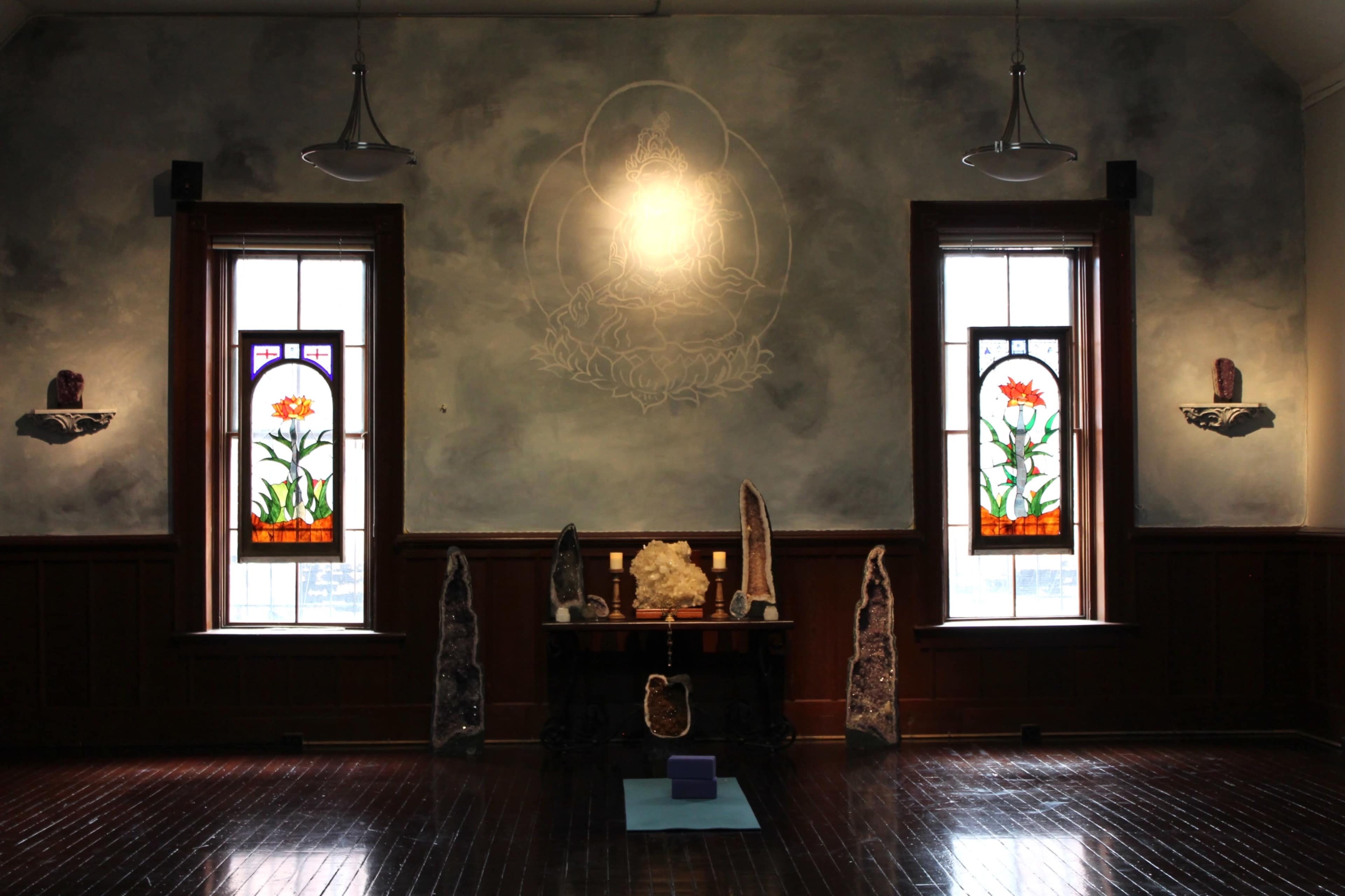A meditation room features two stained glass windows, a central altar with a statue, and large stone formations placed beside the windows.