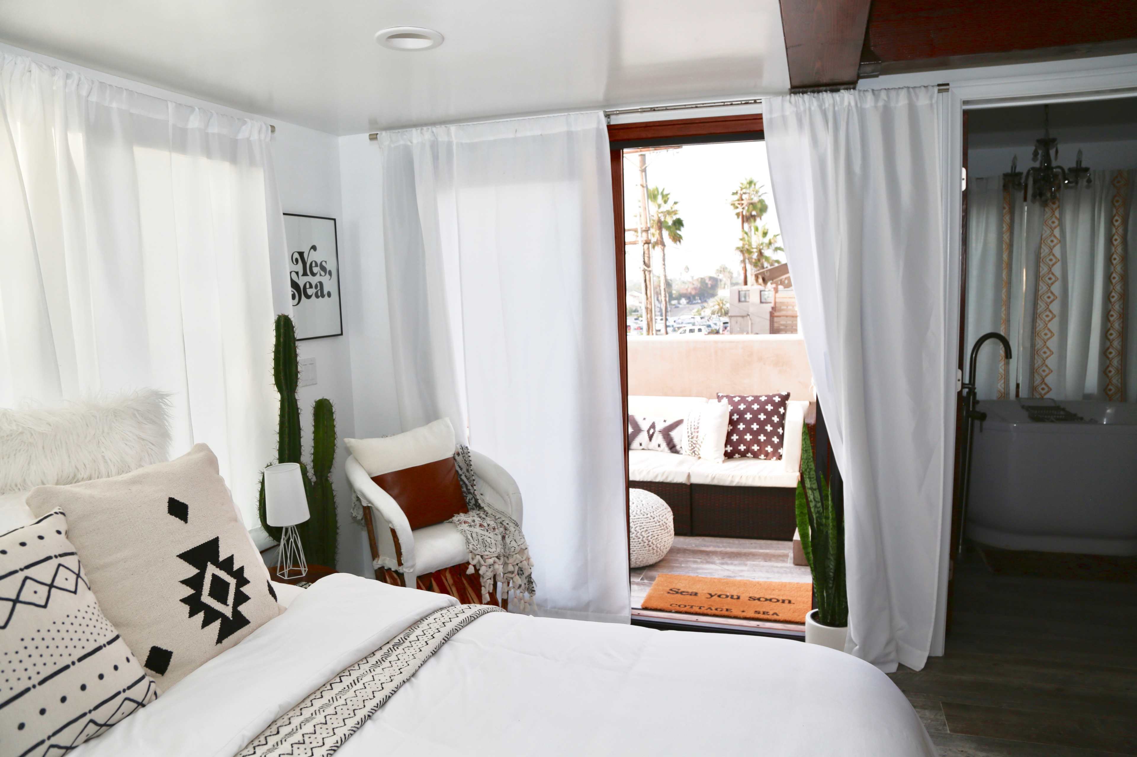 A bright bedroom features a bed with decorative pillows, a white chair, a window opening to a patio, and potted cacti.