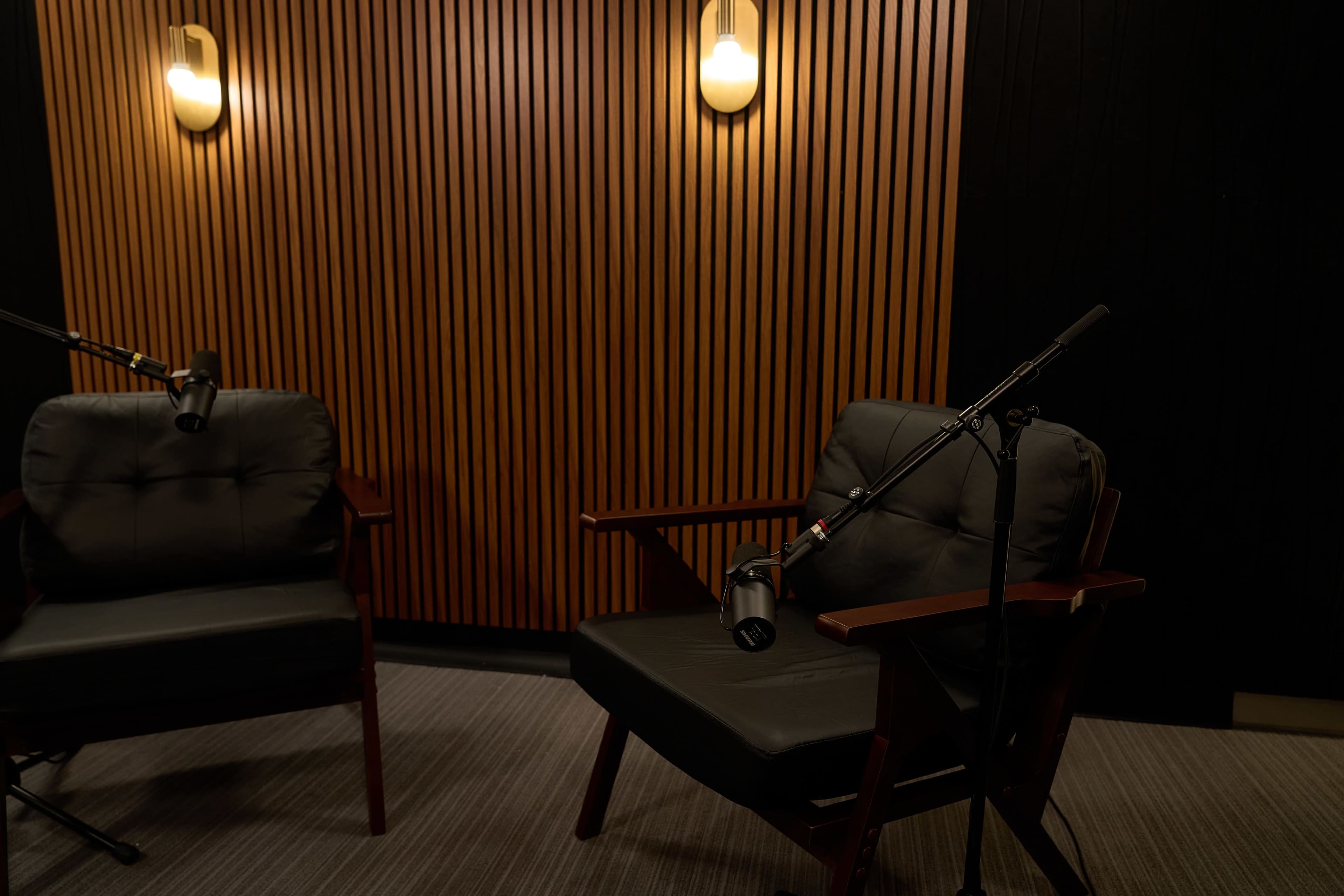 The image shows two black cushioned chairs placed in a room with wooden paneling and microphones positioned over them.