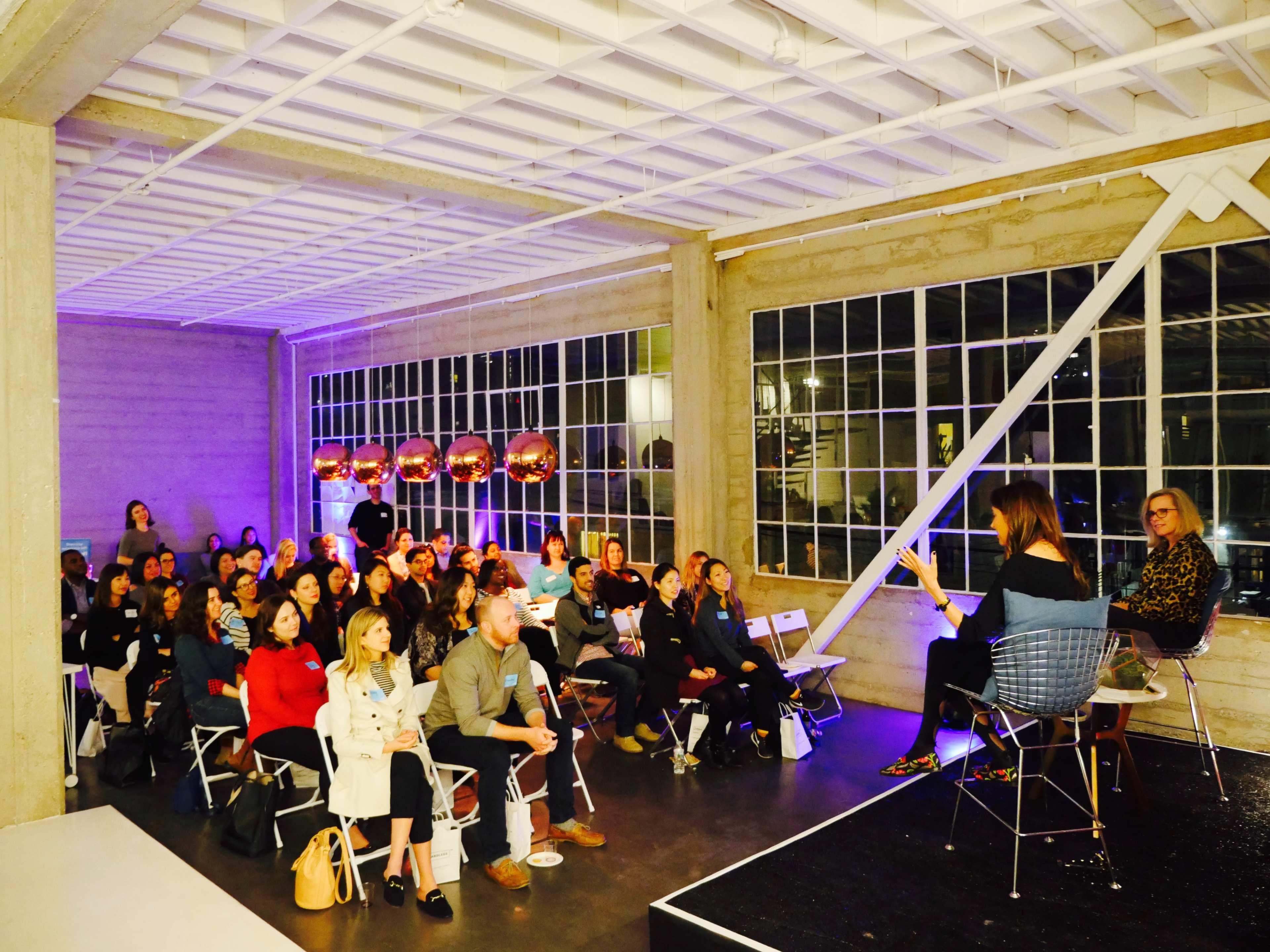 A large audience is seated in a spacious, well-lit venue, listening to speakers on a stage during an event.