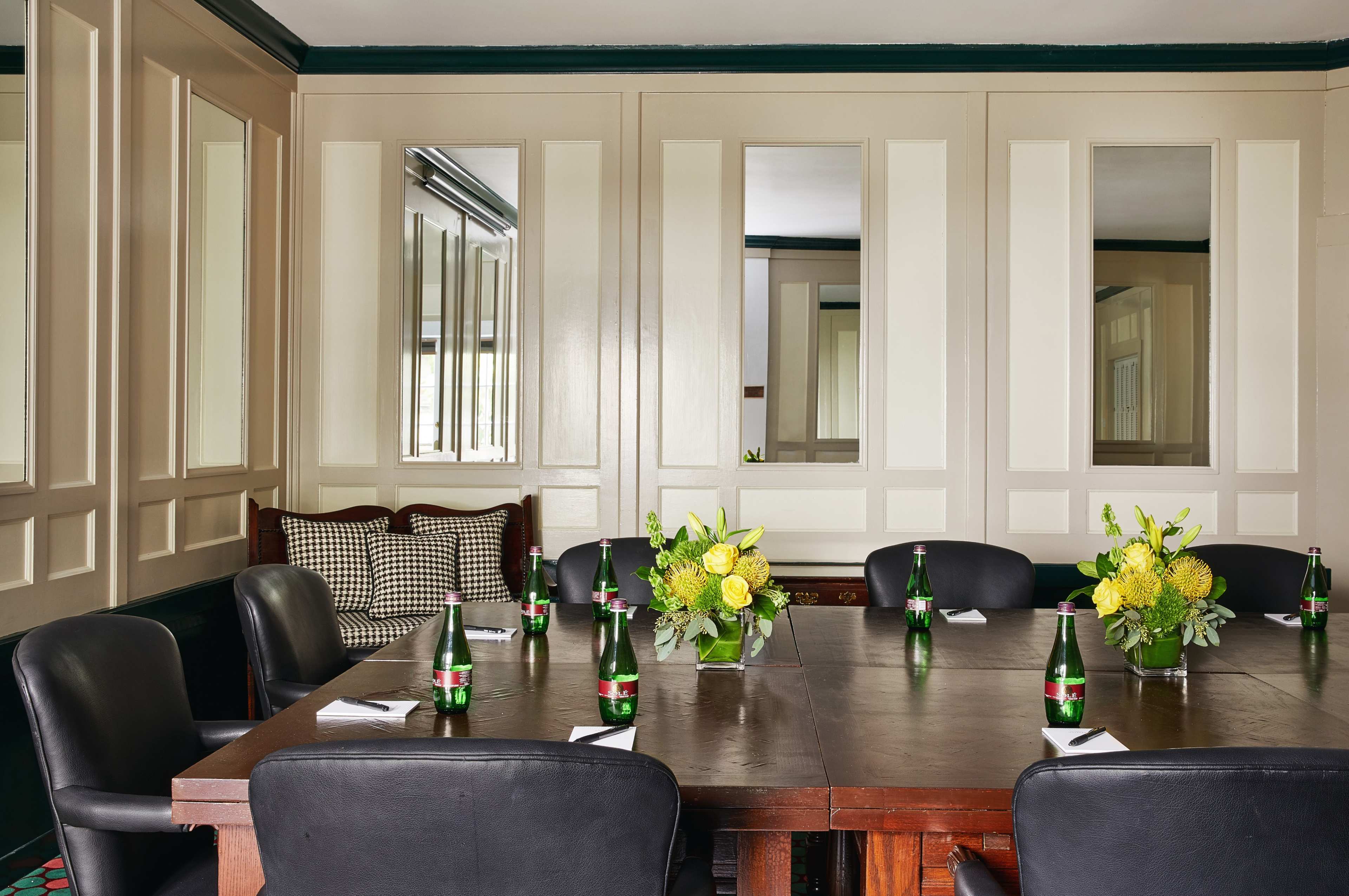 A conference room features a wooden table surrounded by black chairs, decorated with floral arrangements and bottles of sparkling water.