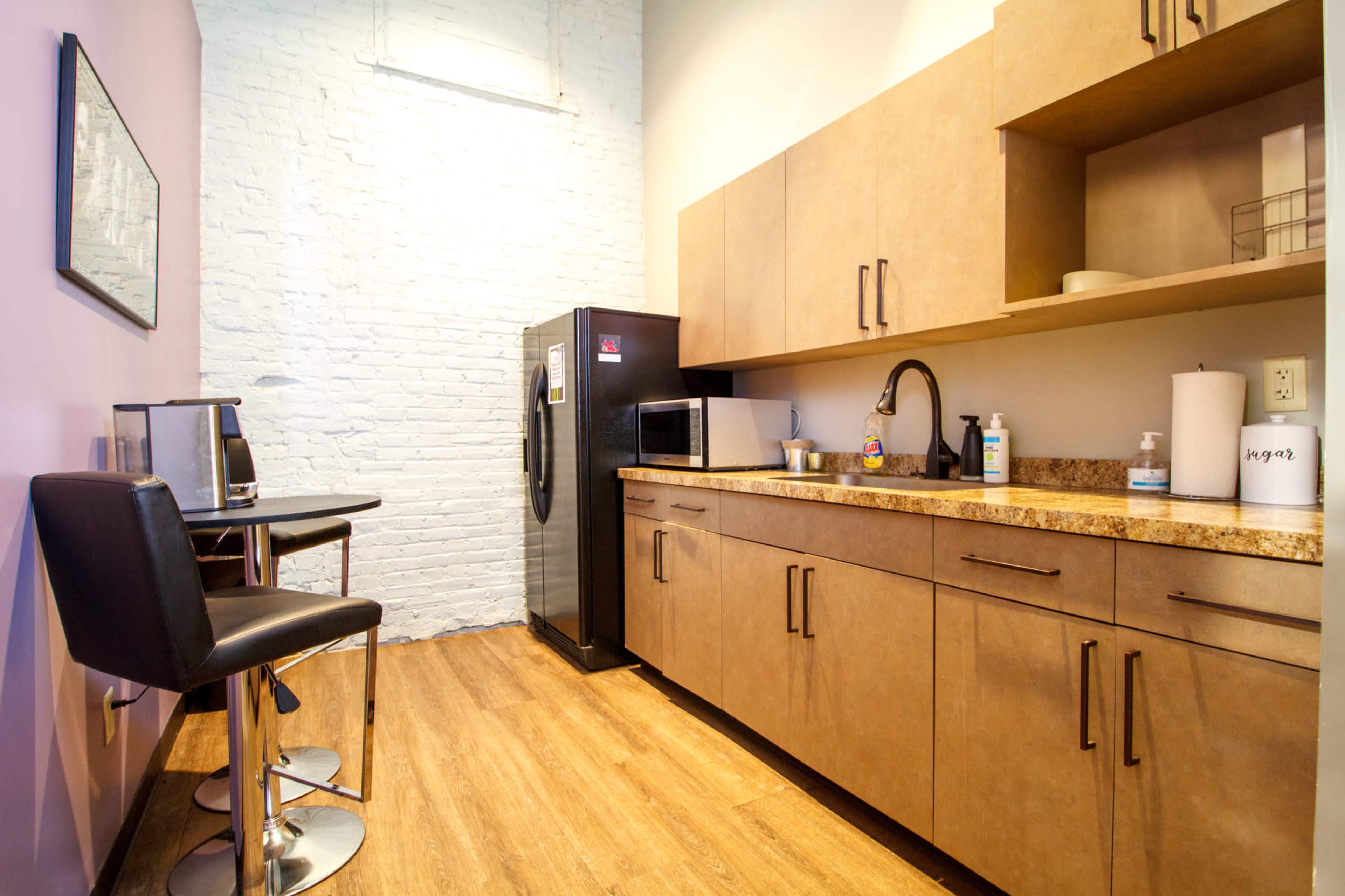 The image shows a small kitchen area with a modern design, featuring a black refrigerator, a countertop, and a two-person table with stylish black chairs.