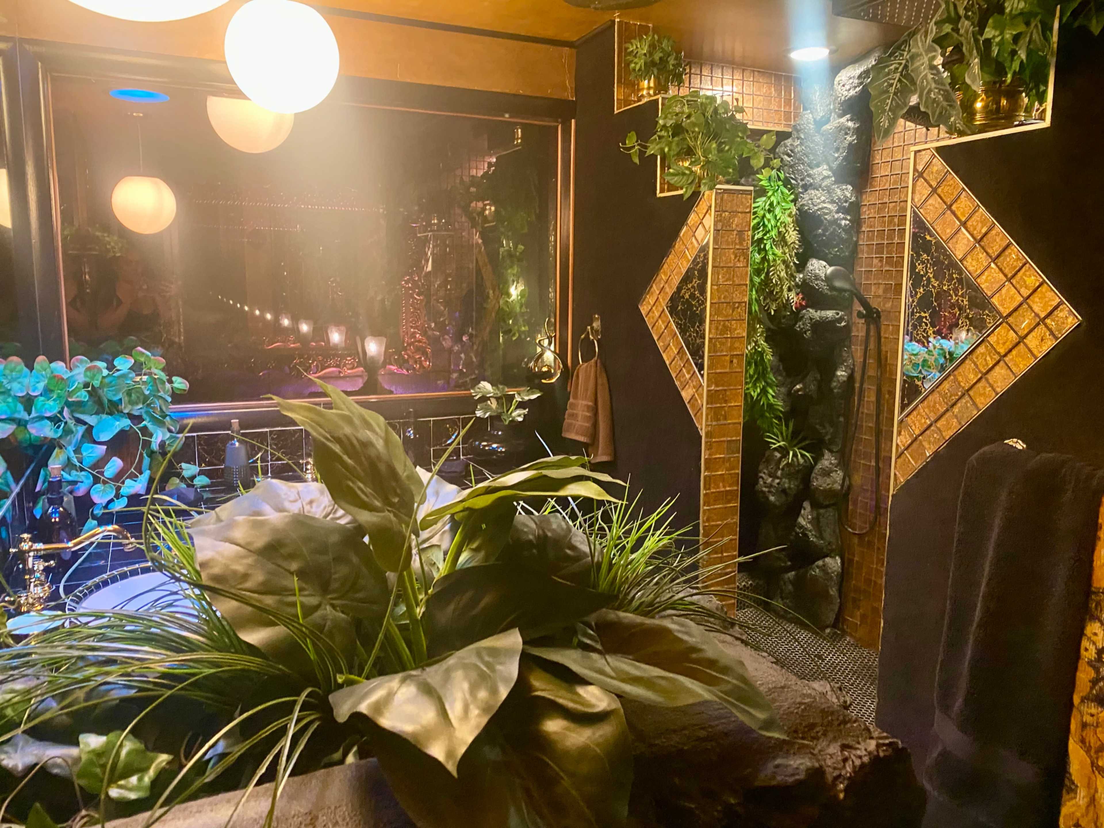 The image shows a modern bathroom with a large stone bathtub surrounded by lush plants, a decorative tiled wall, and a glass shower area.