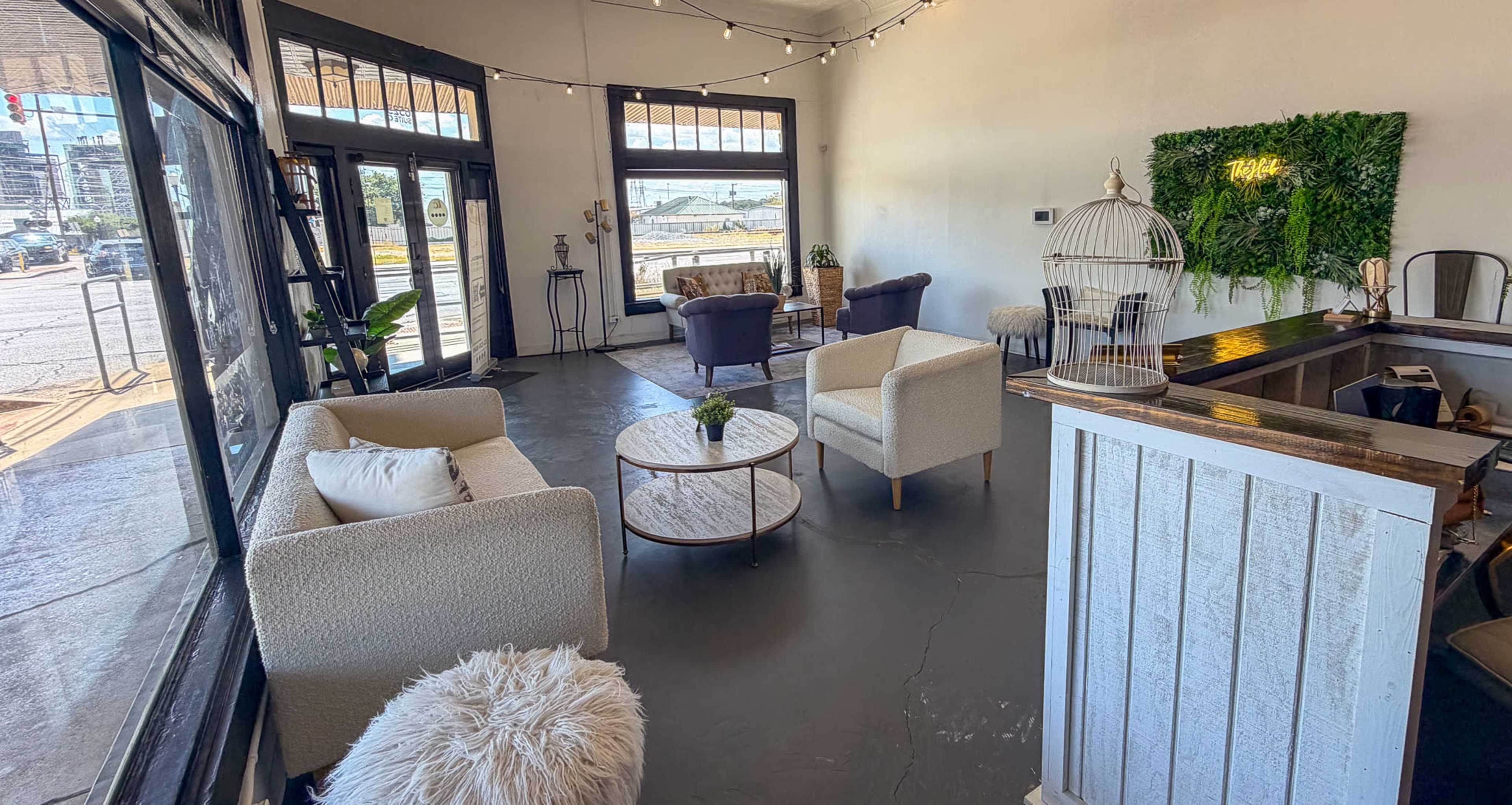The image shows a spacious, well-lit interior of a modern lounge with seating arrangements, including chairs and a small table, and large windows that provide a view of the outside.