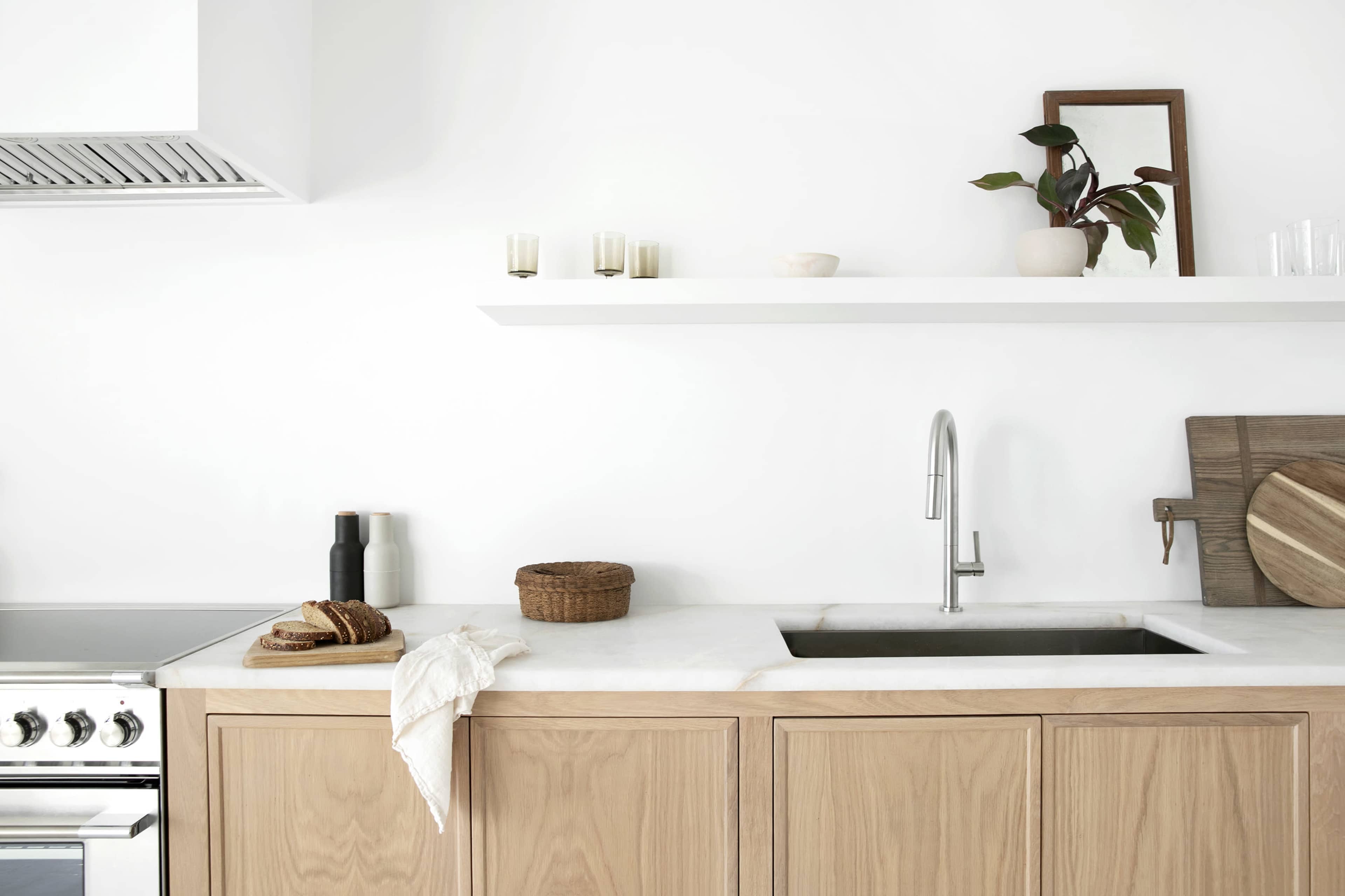 The image shows a modern kitchen with wooden cabinetry, a marble countertop, and minimal decor on open shelves.