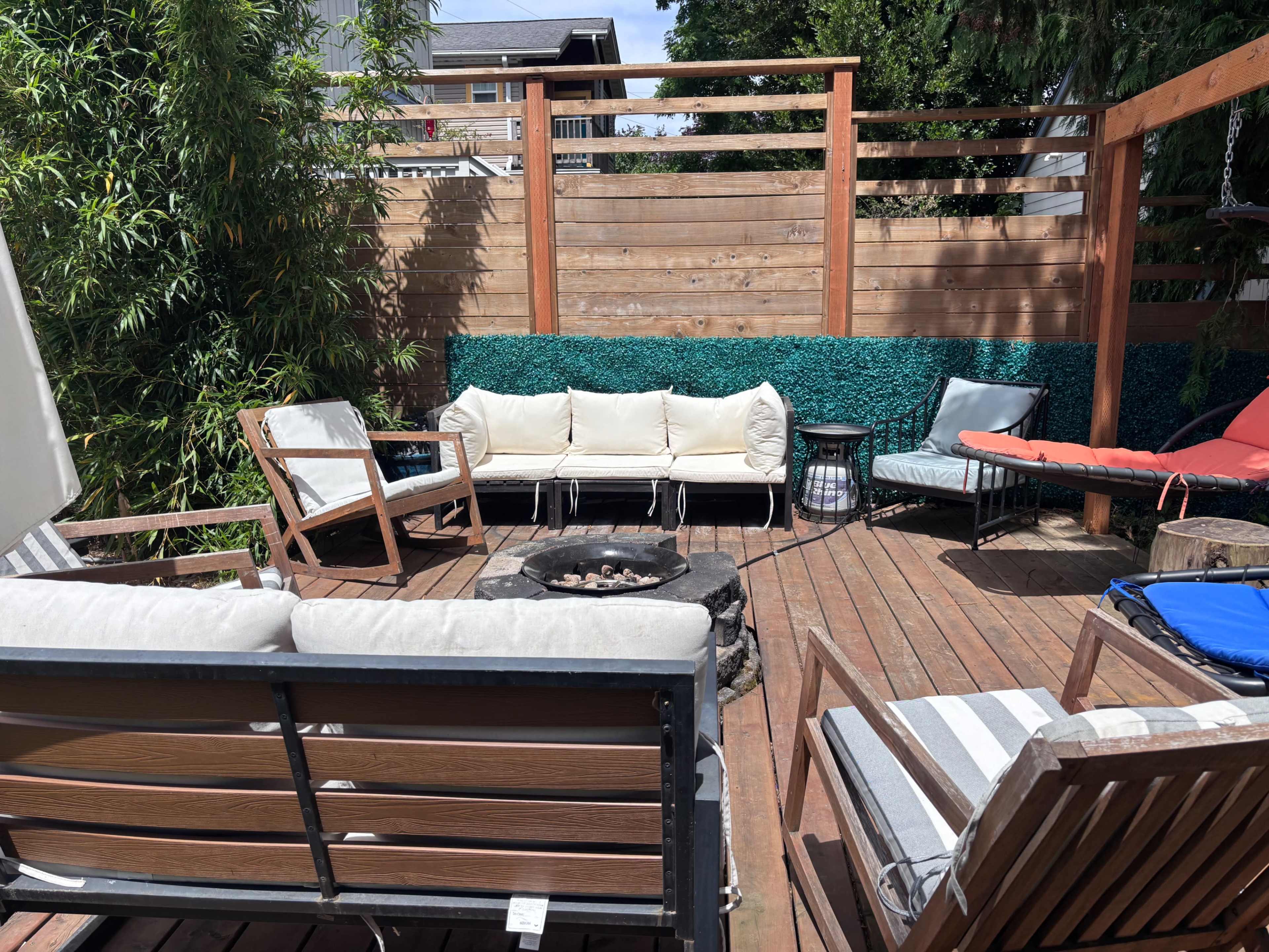 The image shows a wooden patio area with several seating arrangements and a fire pit at its center, surrounded by greenery.