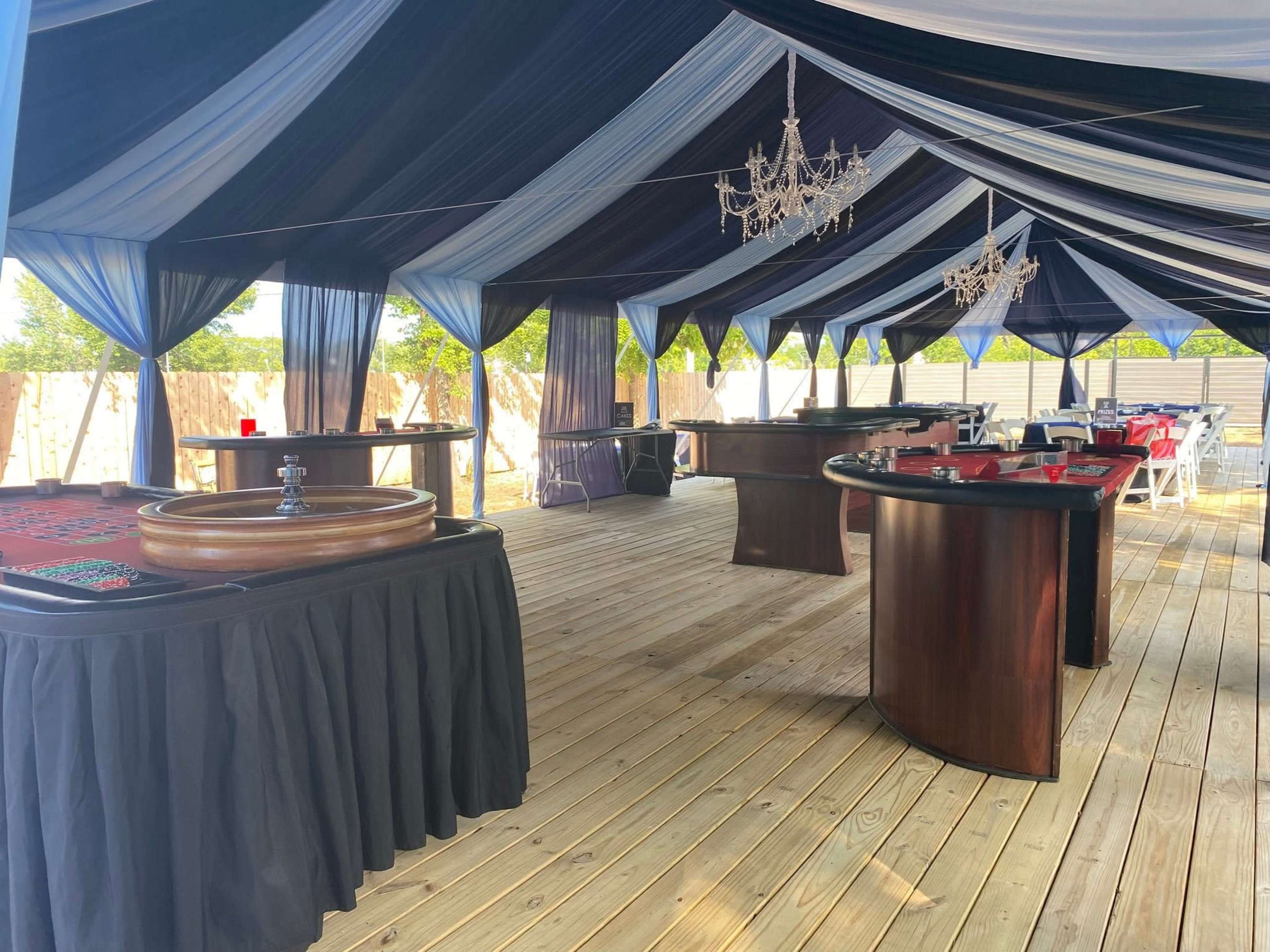The image shows an indoor casino setup with several gaming tables and a large, decorative tent featuring draped blue and black fabric.