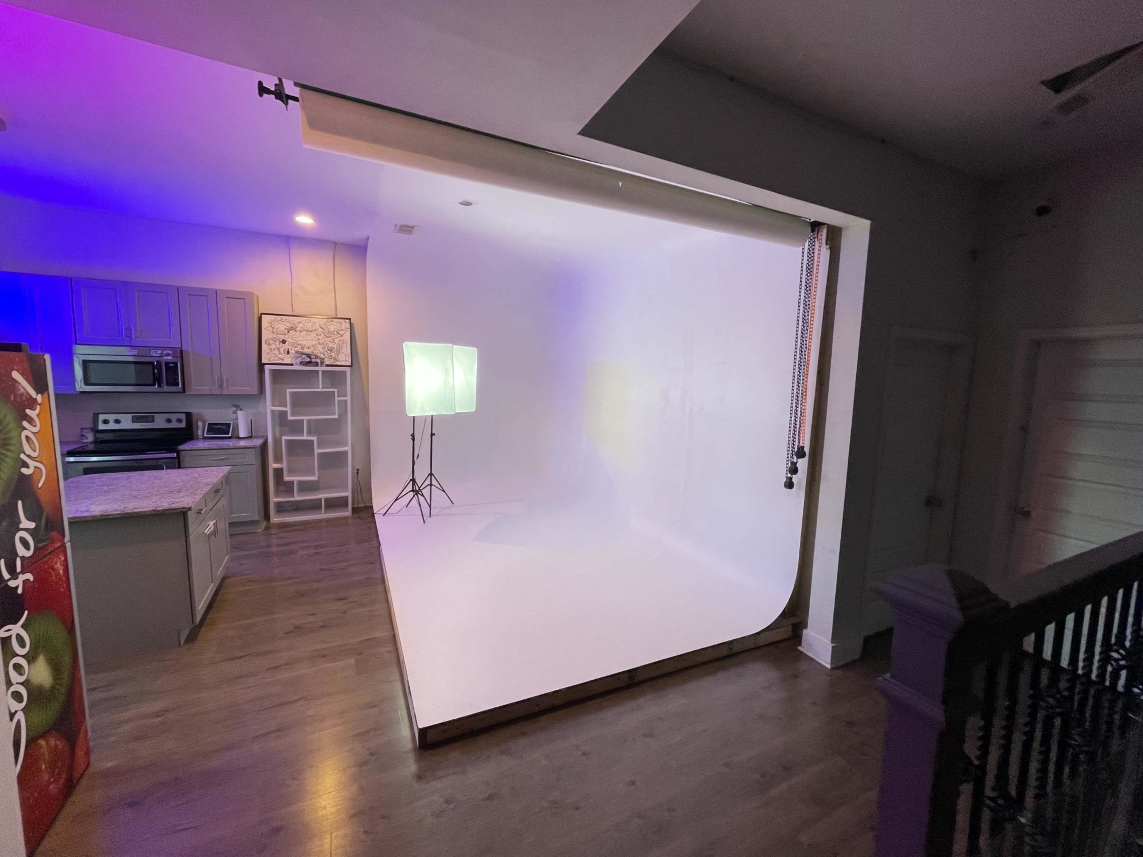 A spacious room featuring a kitchen on one side and a white backdrop setup with lighting equipment on the other.
