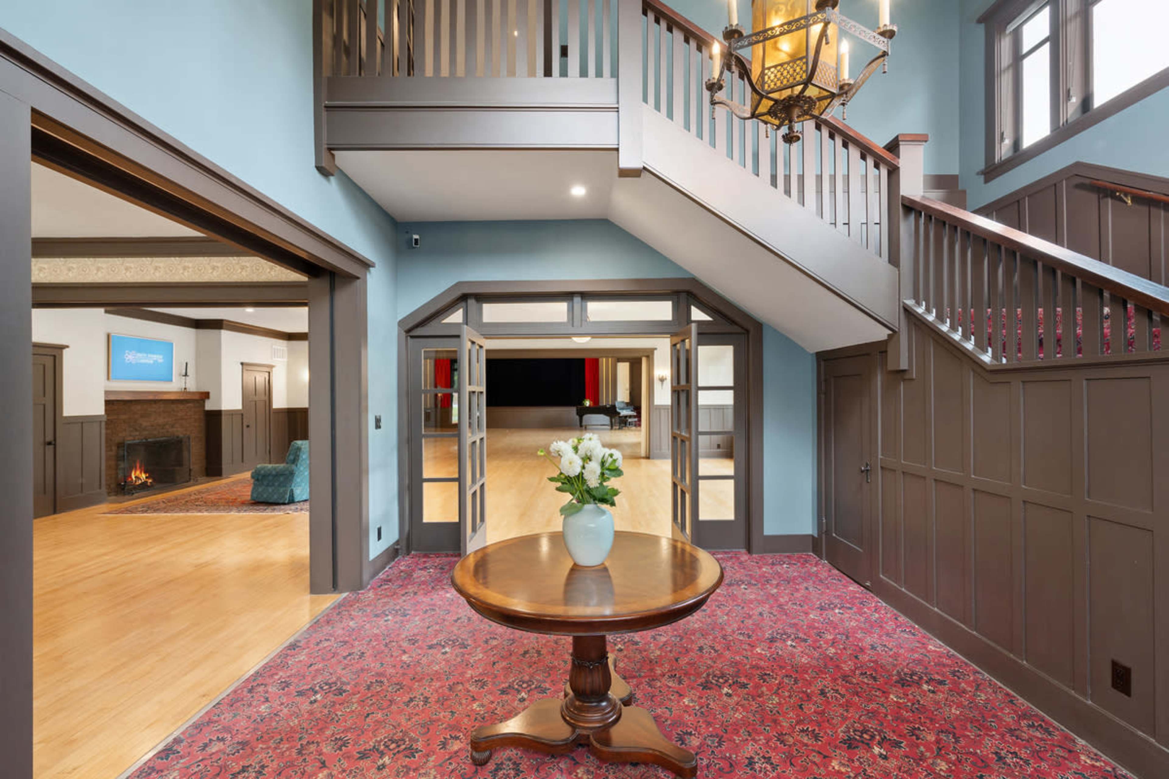 The image shows a large foyer with a round wooden table at its center, a staircase leading to an upper floor, and entranceways to adjacent rooms.