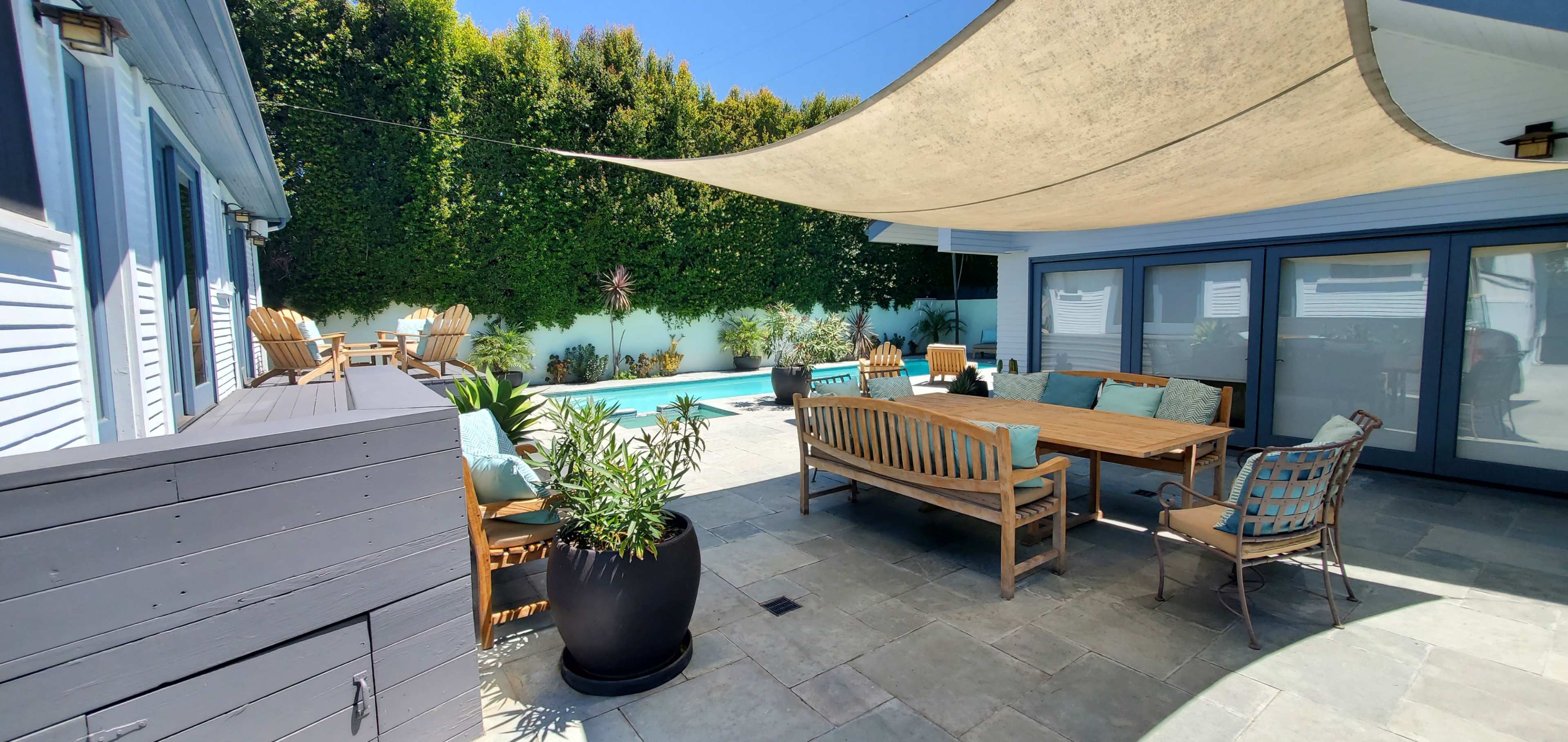 A patio area features a large table surrounded by wooden chairs and a swimming pool in the background, with shade provided by a large canvas awning.