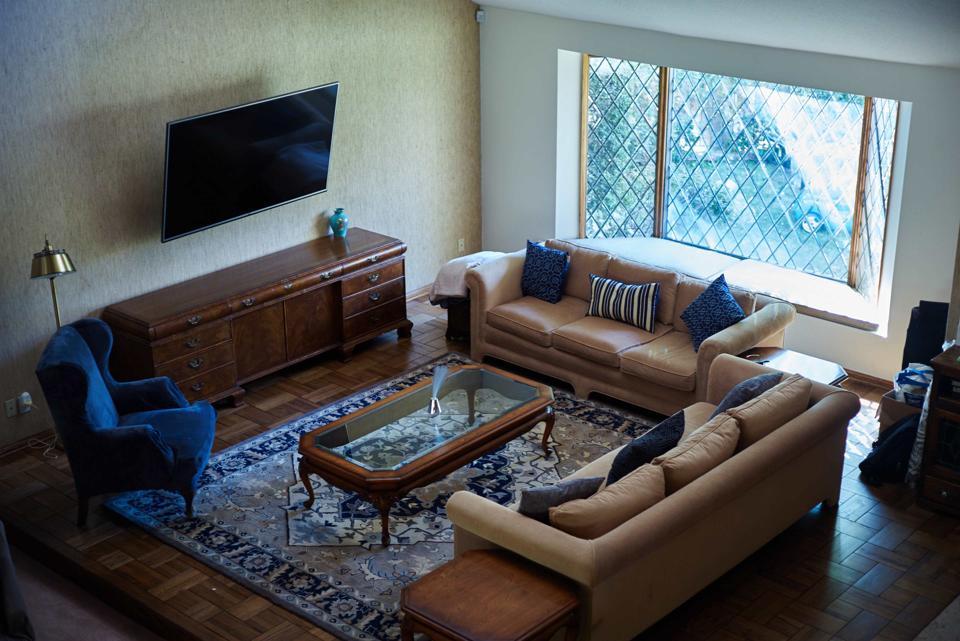 A spacious living room features a television mounted on the wall, a wooden sideboard, two sofas, an armchair, and a large window with a distinctive grid design.