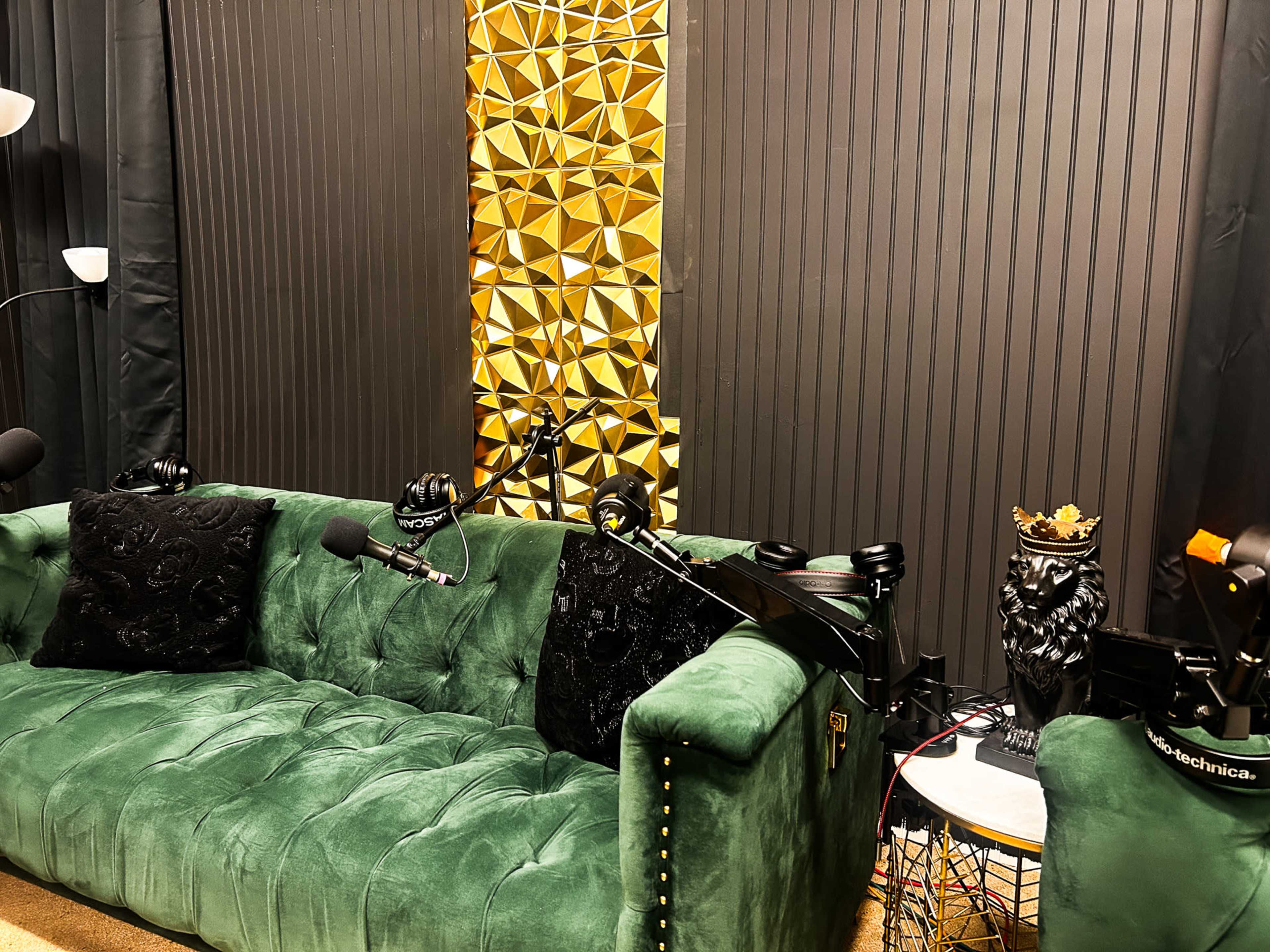 The image shows a green tufted couch placed against a black wall with a gold geometric panel, accompanied by microphones and a decorative lion statue.