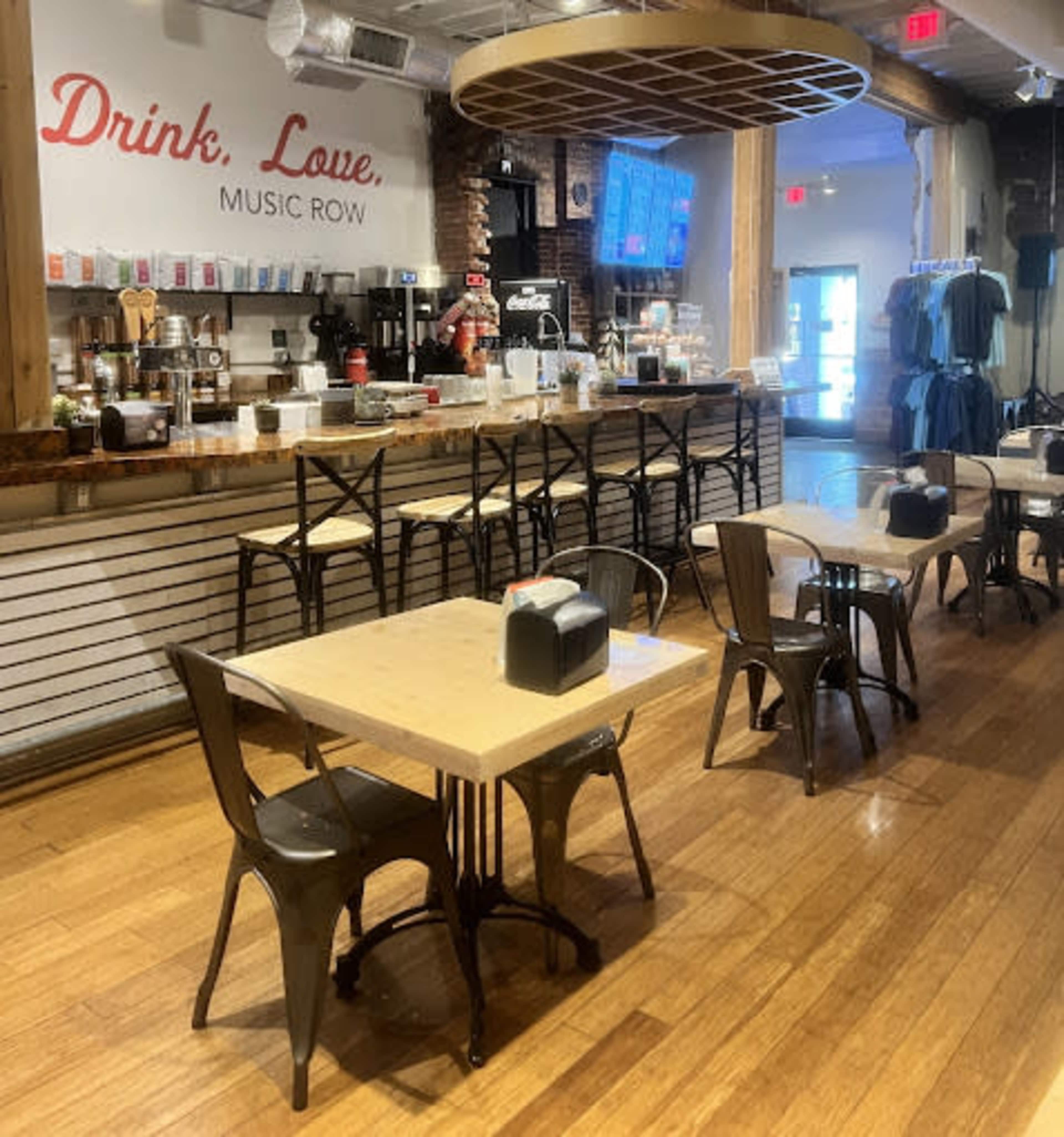 The image shows a modern café interior with several wooden tables and metal chairs, a coffee counter lined with beverage equipment, and merchandise displayed on the walls.
