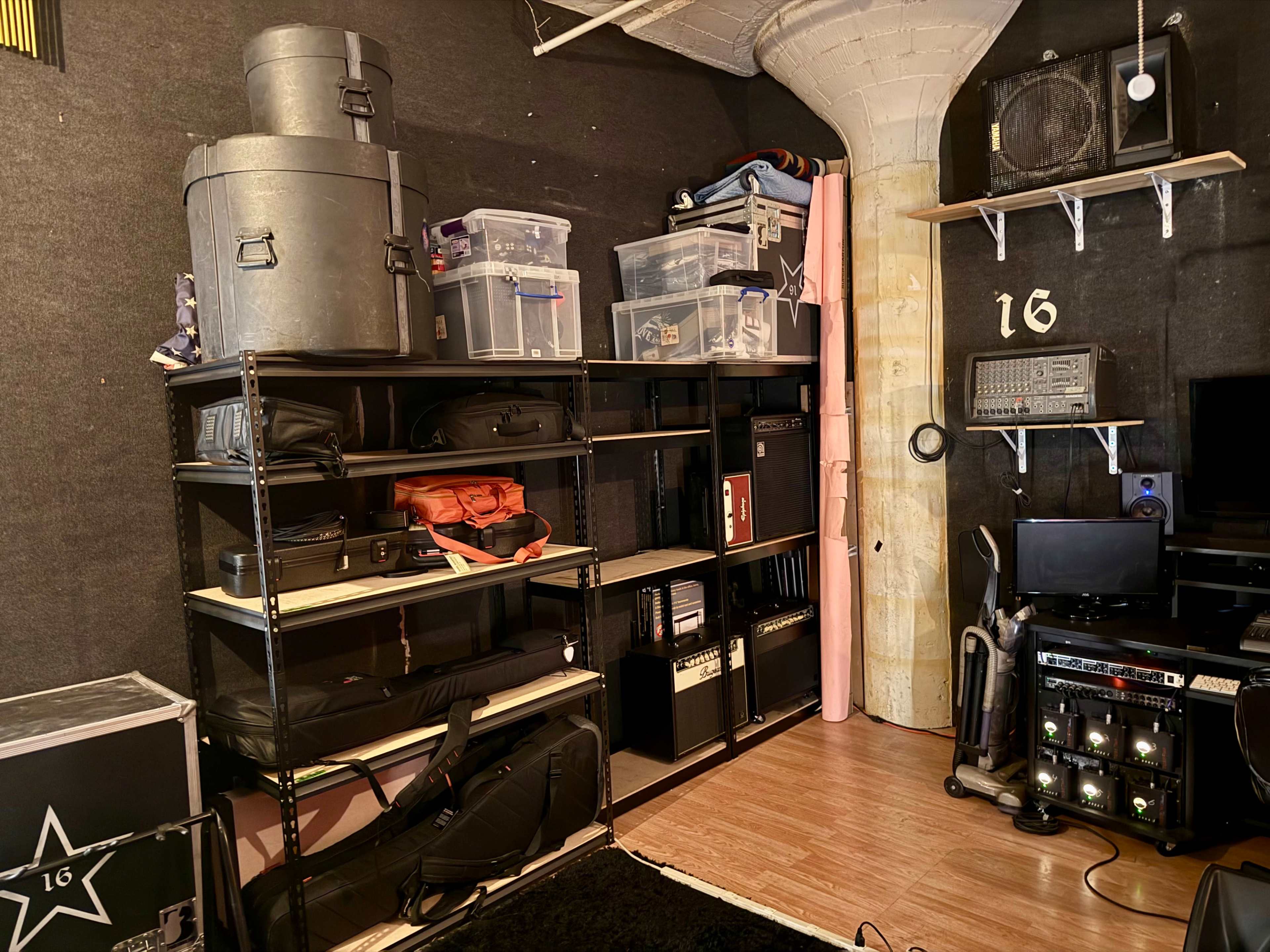 The image shows a room with shelves containing various storage containers, musical equipment, and a television setup against a textured wall.