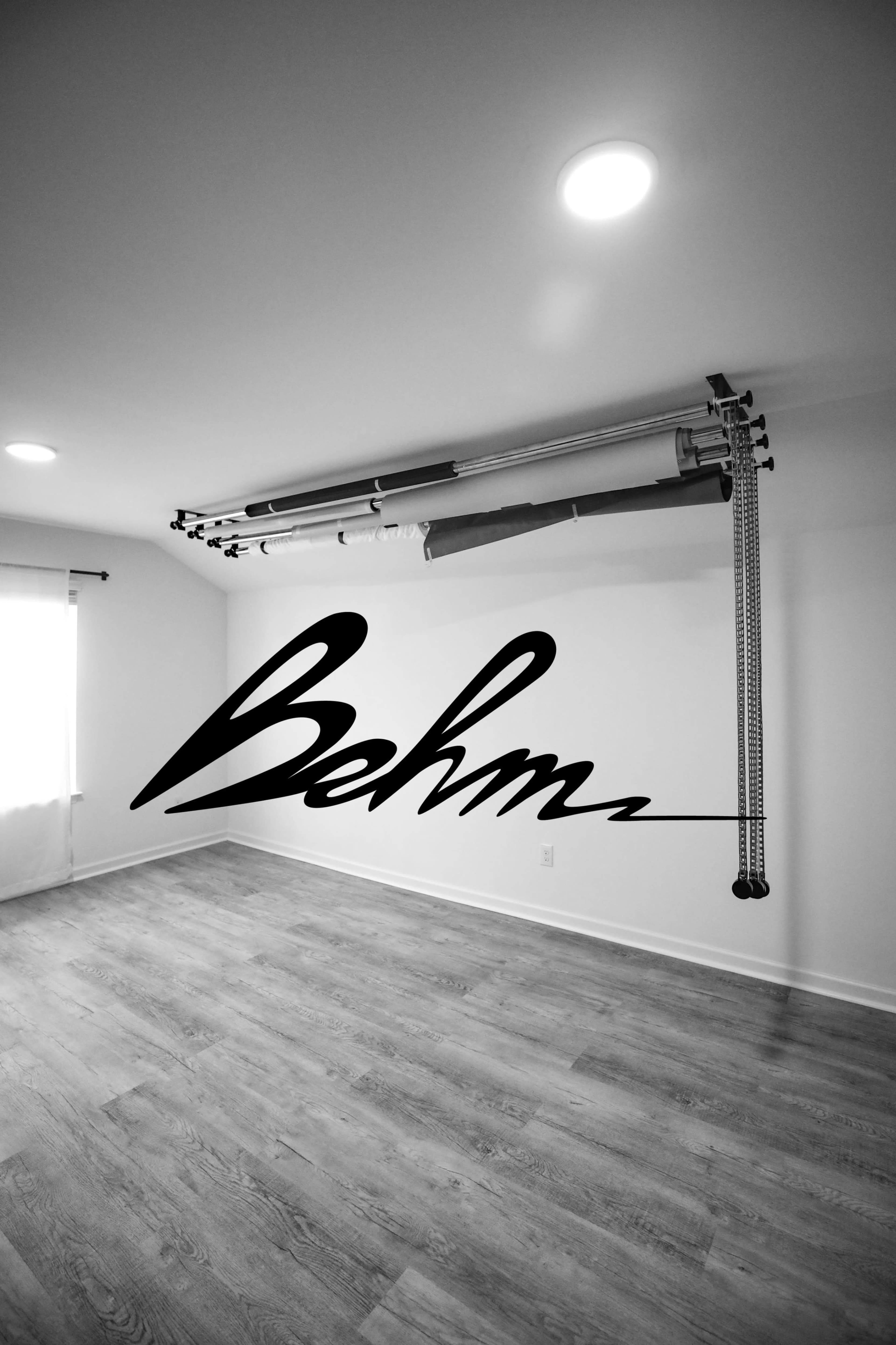 The image shows a spacious, empty room with a large overhead curtain rod and a stylized "Behm" graphic on the wall.