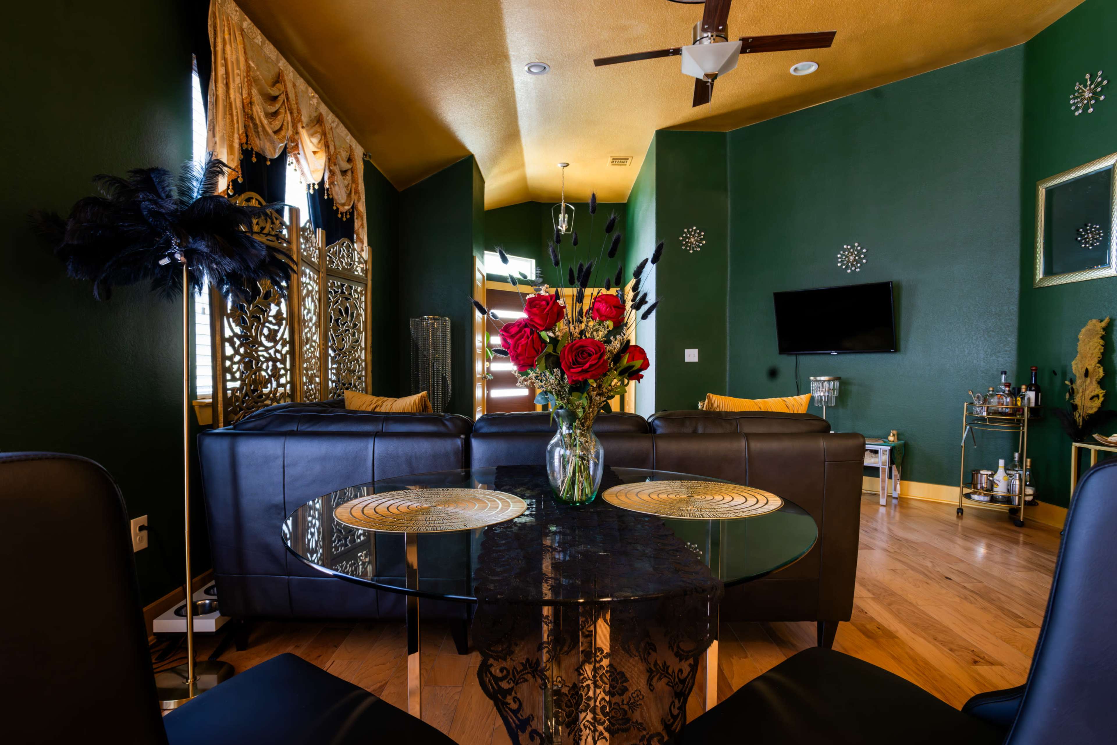 A stylish living area with a glass dining table, a vase of red roses, and dark leather couches against a green wall.