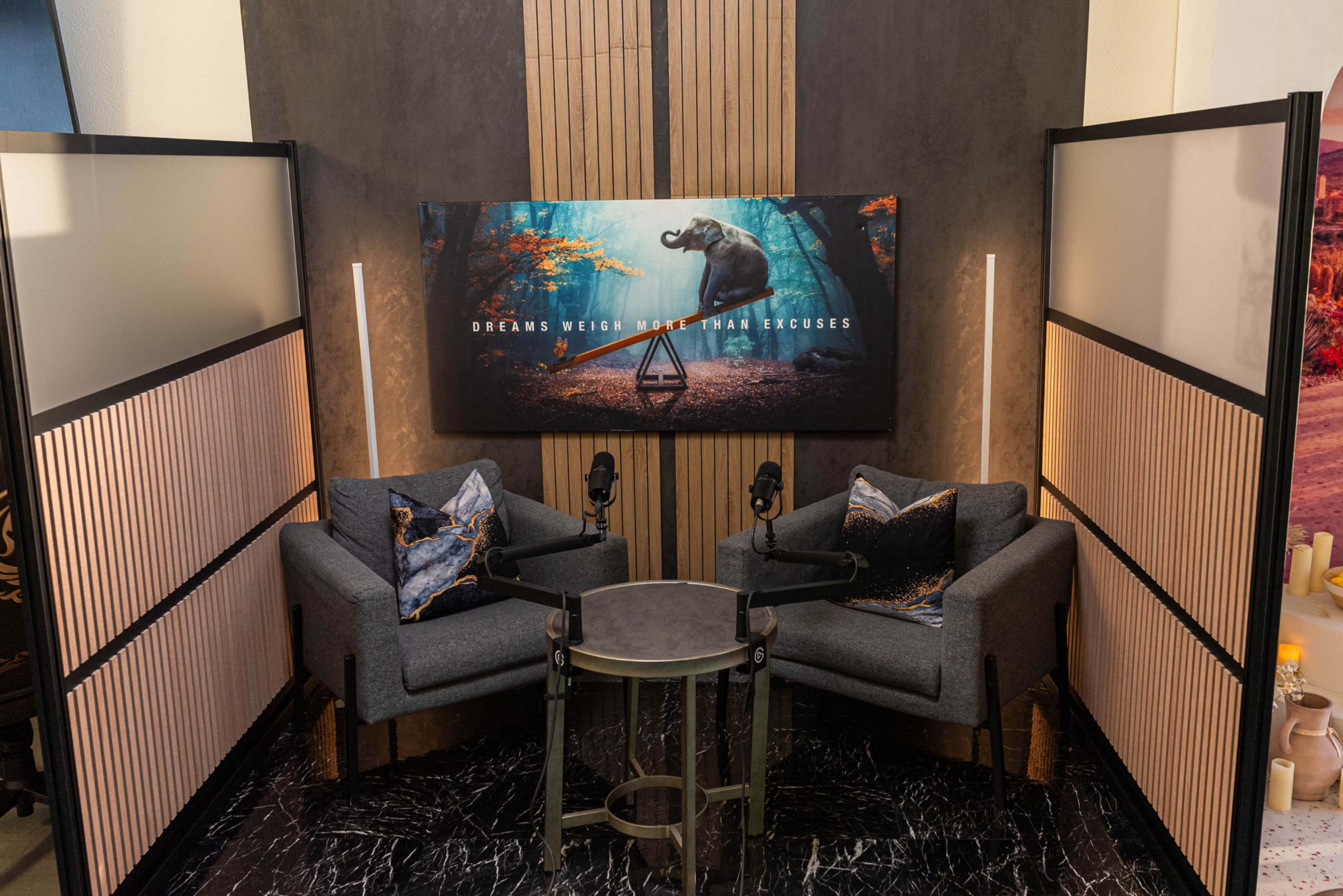 The image shows a modern conversation area with two grey chairs, a small table, and a decorative backdrop featuring an elephant and the phrase "DREAMS WEIGH MORE THAN EXCUSES."