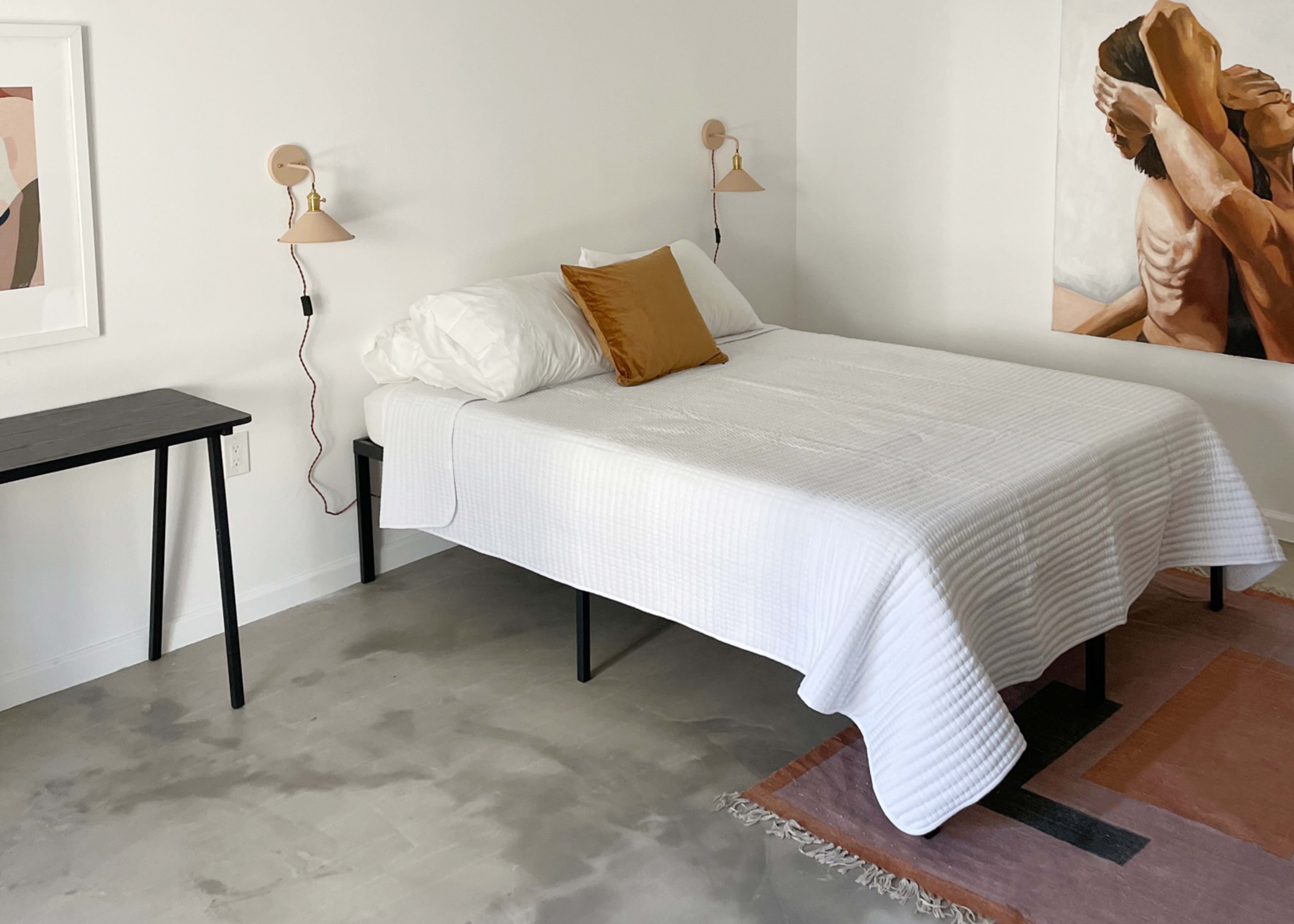 A minimalistic bedroom featuring a bed with a white blanket and a decorative pillow, accompanied by two wall-mounted lamps and a small black table against the wall.