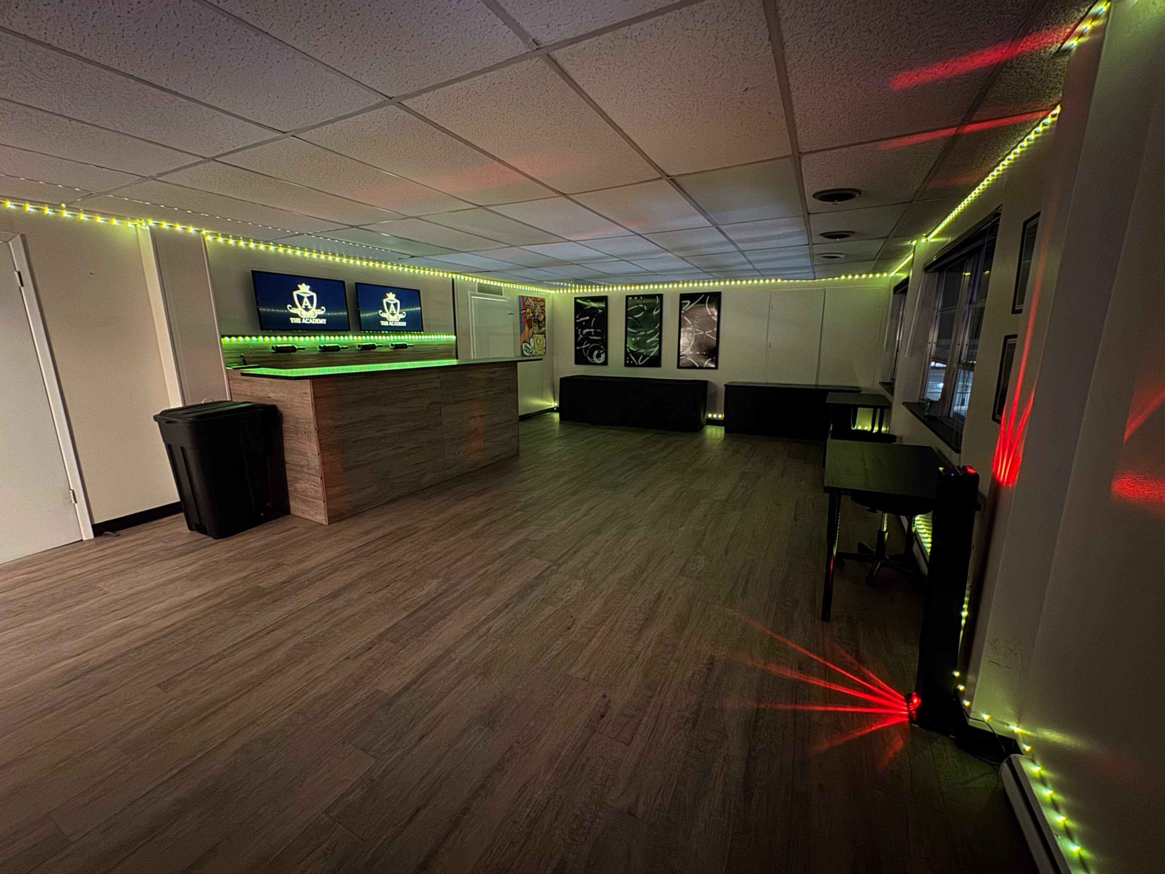 The image shows a modern, empty room with a reception counter, illuminated by green and red LED lights along the walls, featuring artwork and a trash bin.