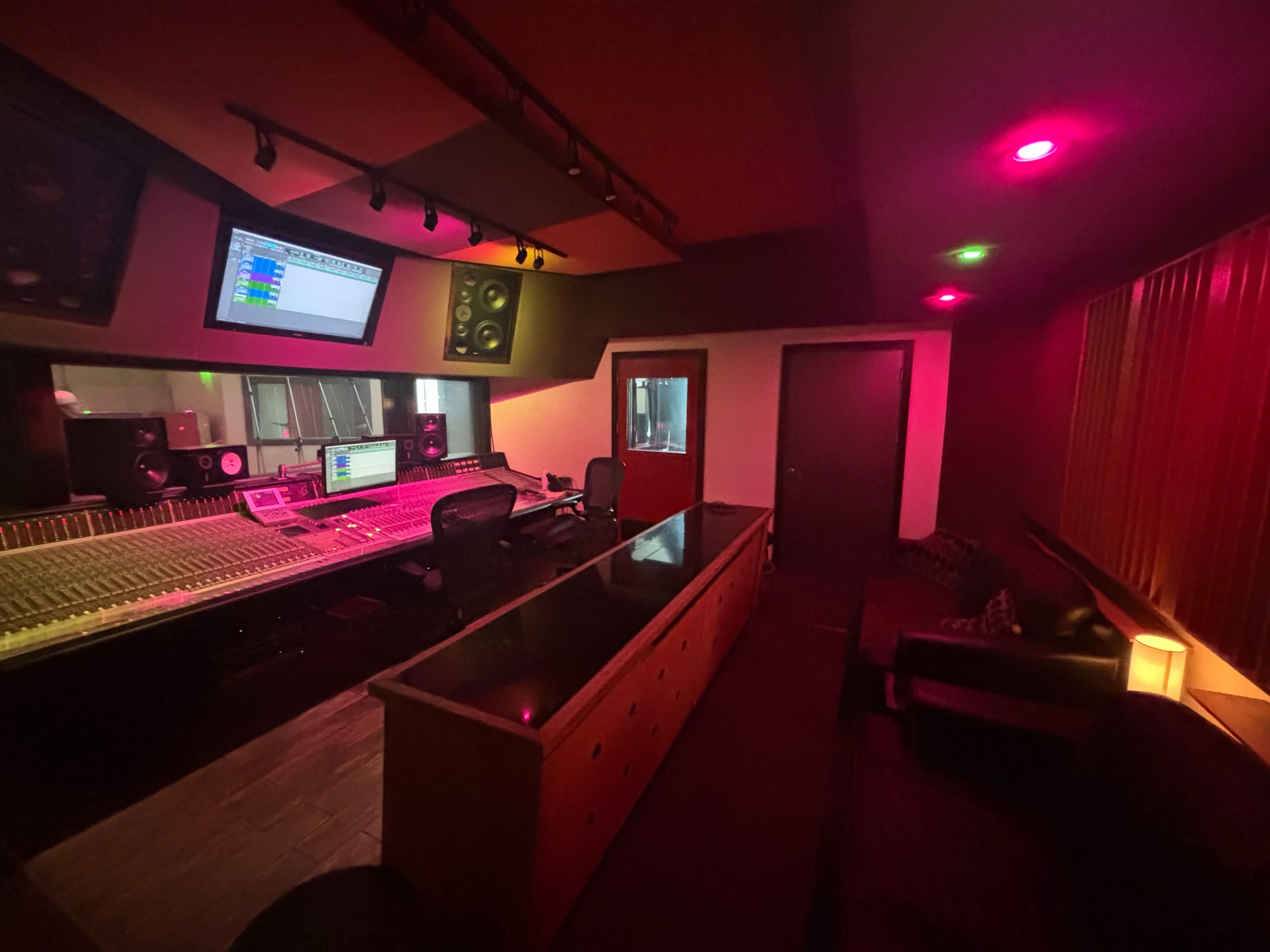 The image shows a music recording studio featuring a large mixing console, several speakers, and a control room with colored lighting.