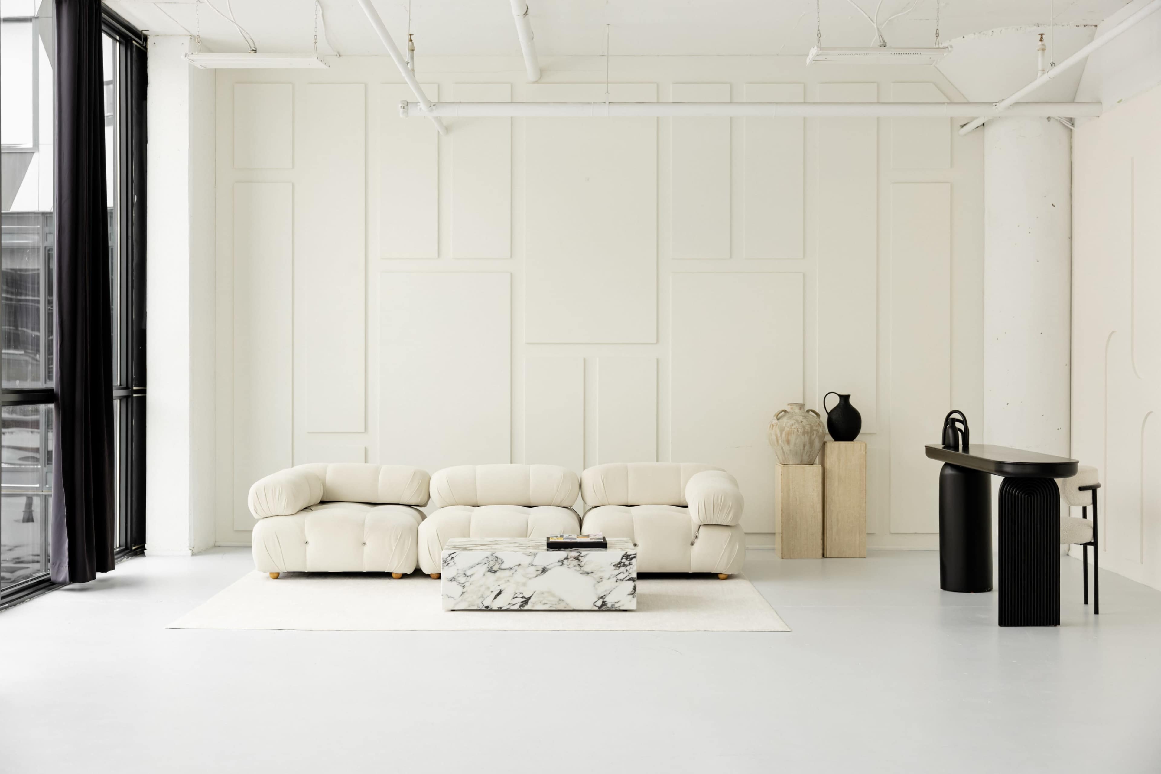 The image features a minimalist living room with a light-colored sectional sofa, a marble coffee table, a black console table, and large windows with sheer curtains.