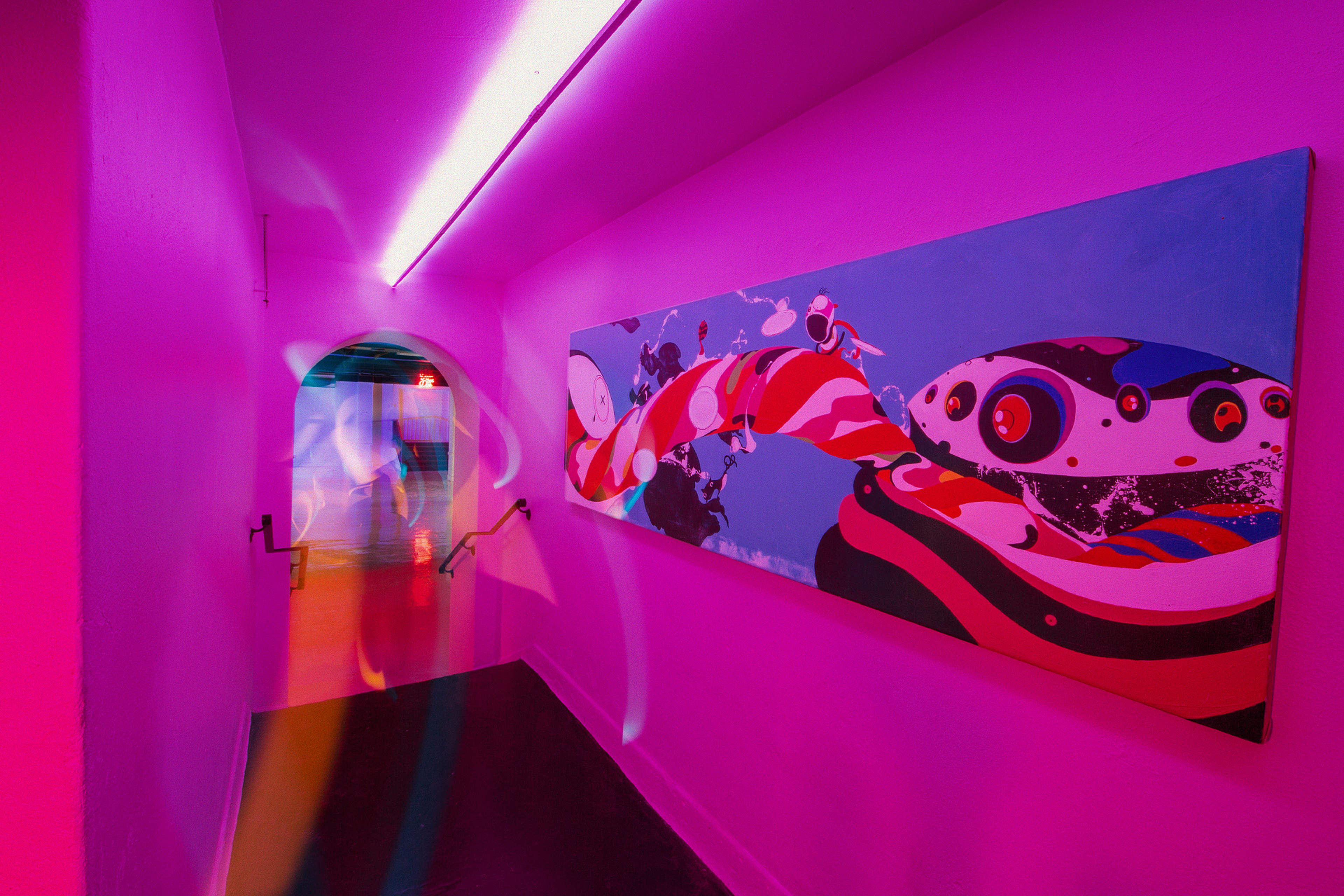 A brightly lit corridor with purple walls features a large colorful mural and a staircase at the end.