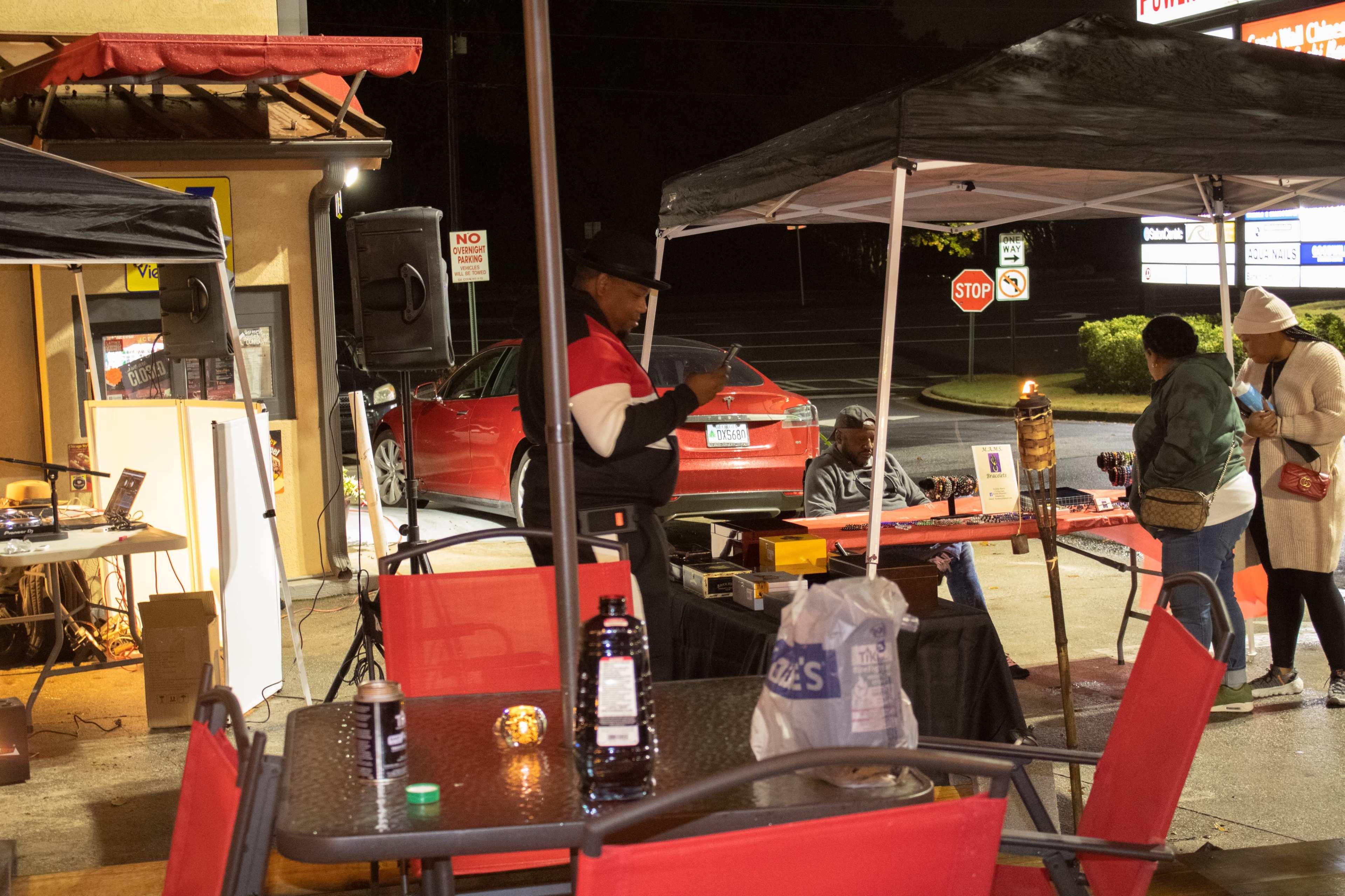 A musician plays at night under a tent near a parking lot, while a small crowd gathers around tables with a red car and signage visible in the background.