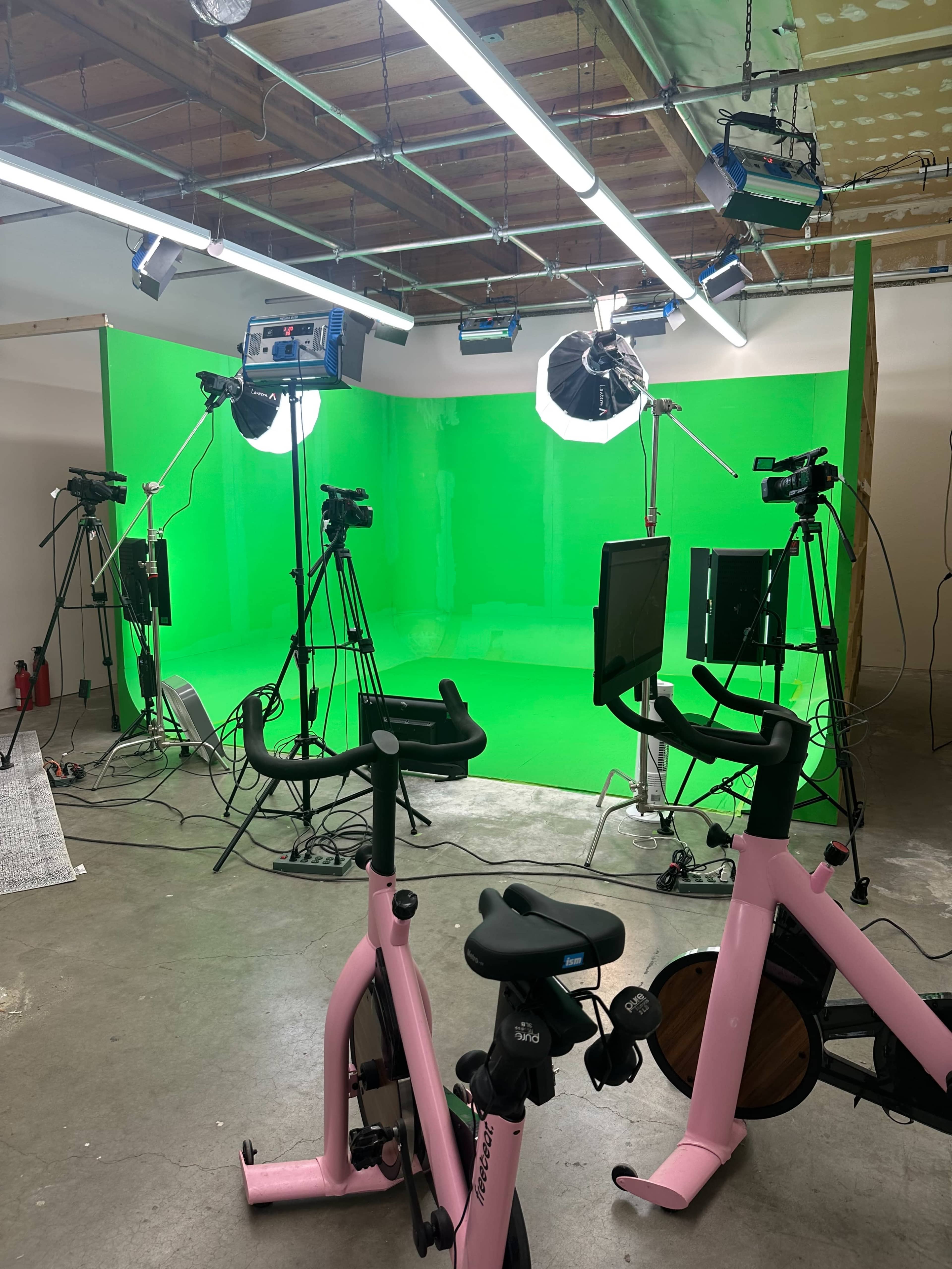 A studio setup with pink exercise bikes in the foreground and multiple cameras, lights, and a green backdrop in the background.