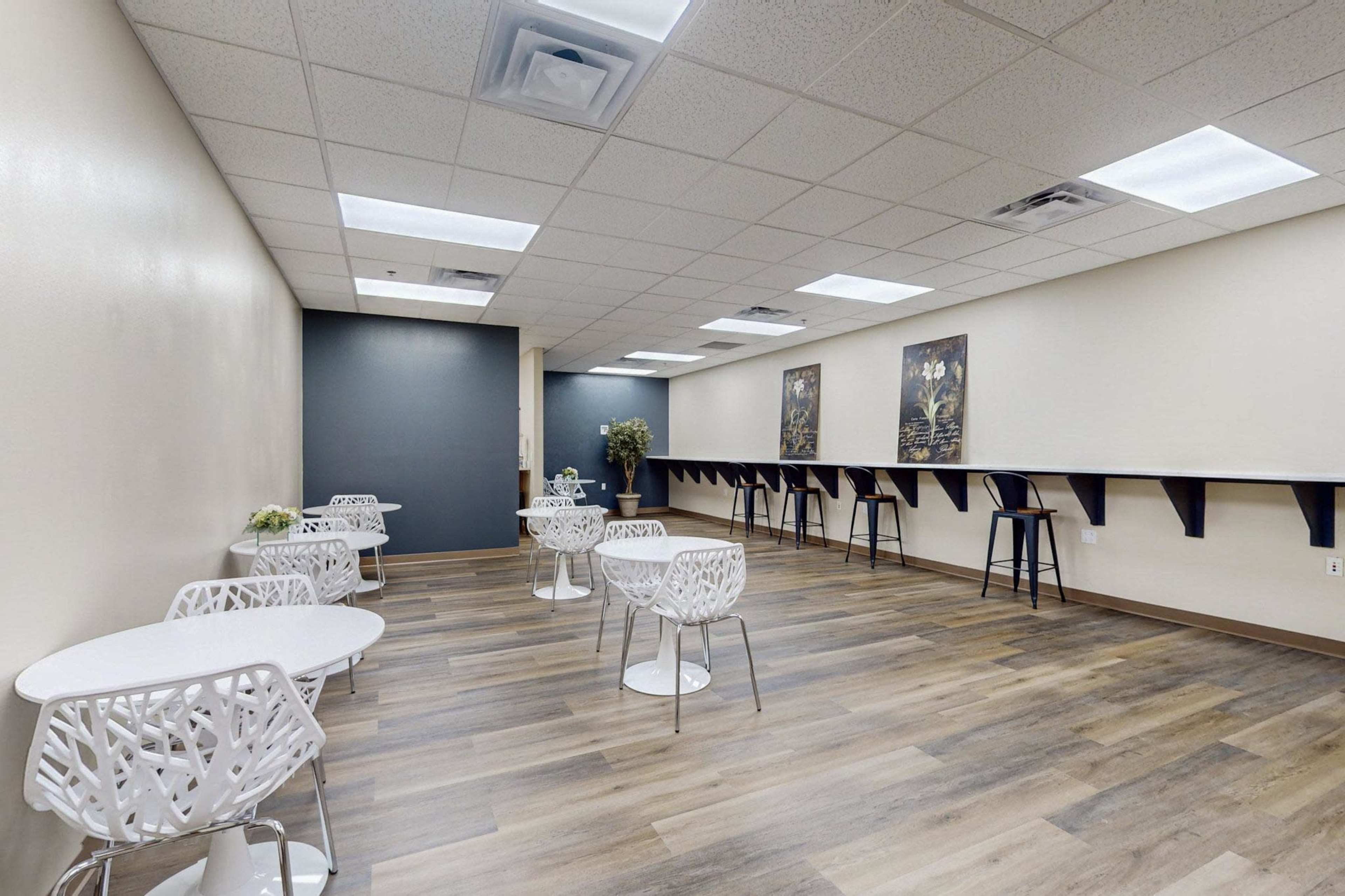 A spacious, light-colored room with white tables and chairs arranged in small groups, featuring a dark accent wall and artwork on the opposite side.