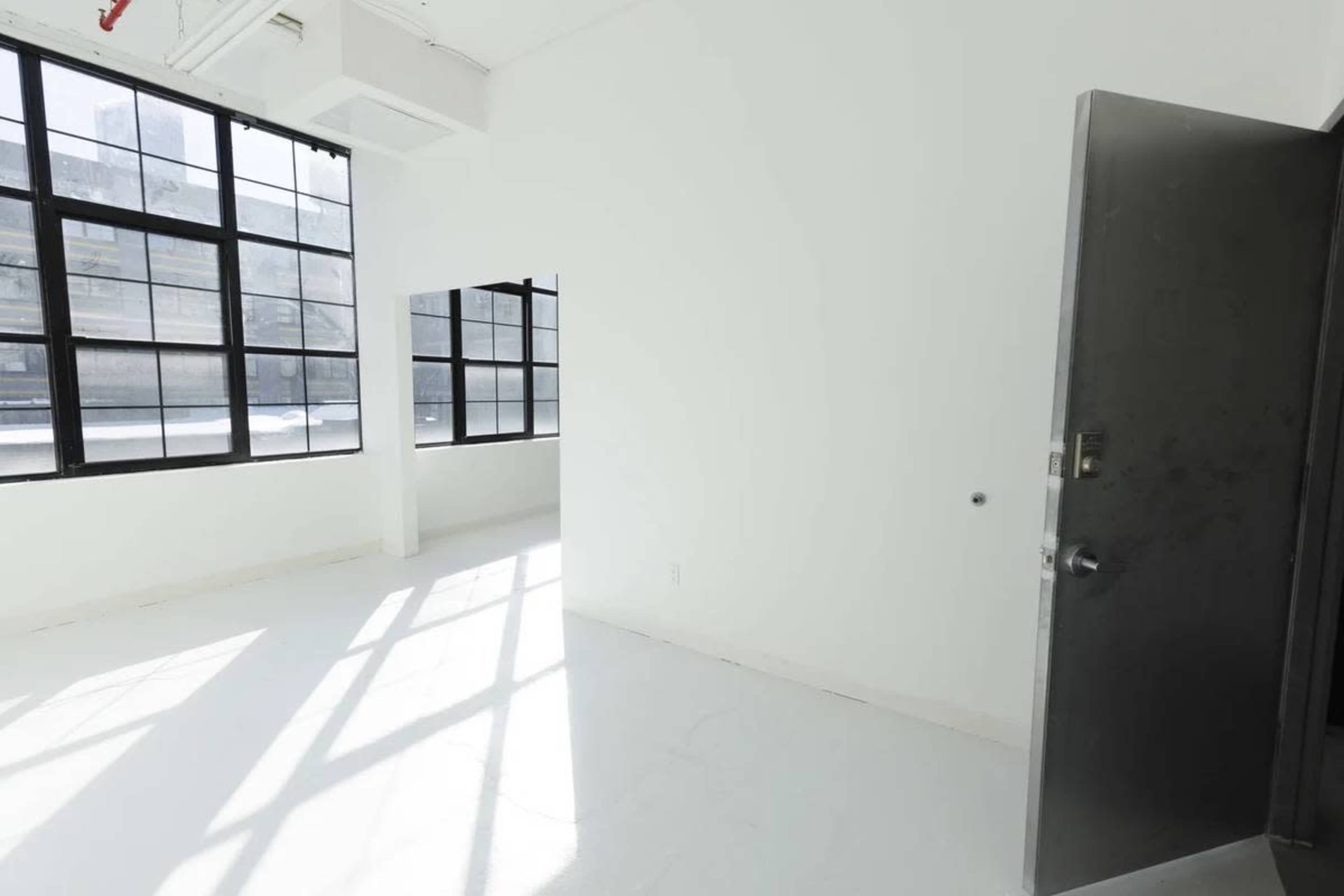 The image shows a bright, spacious room with large windows, a door on the right, and a minimalistic design featuring white walls and a white floor.
