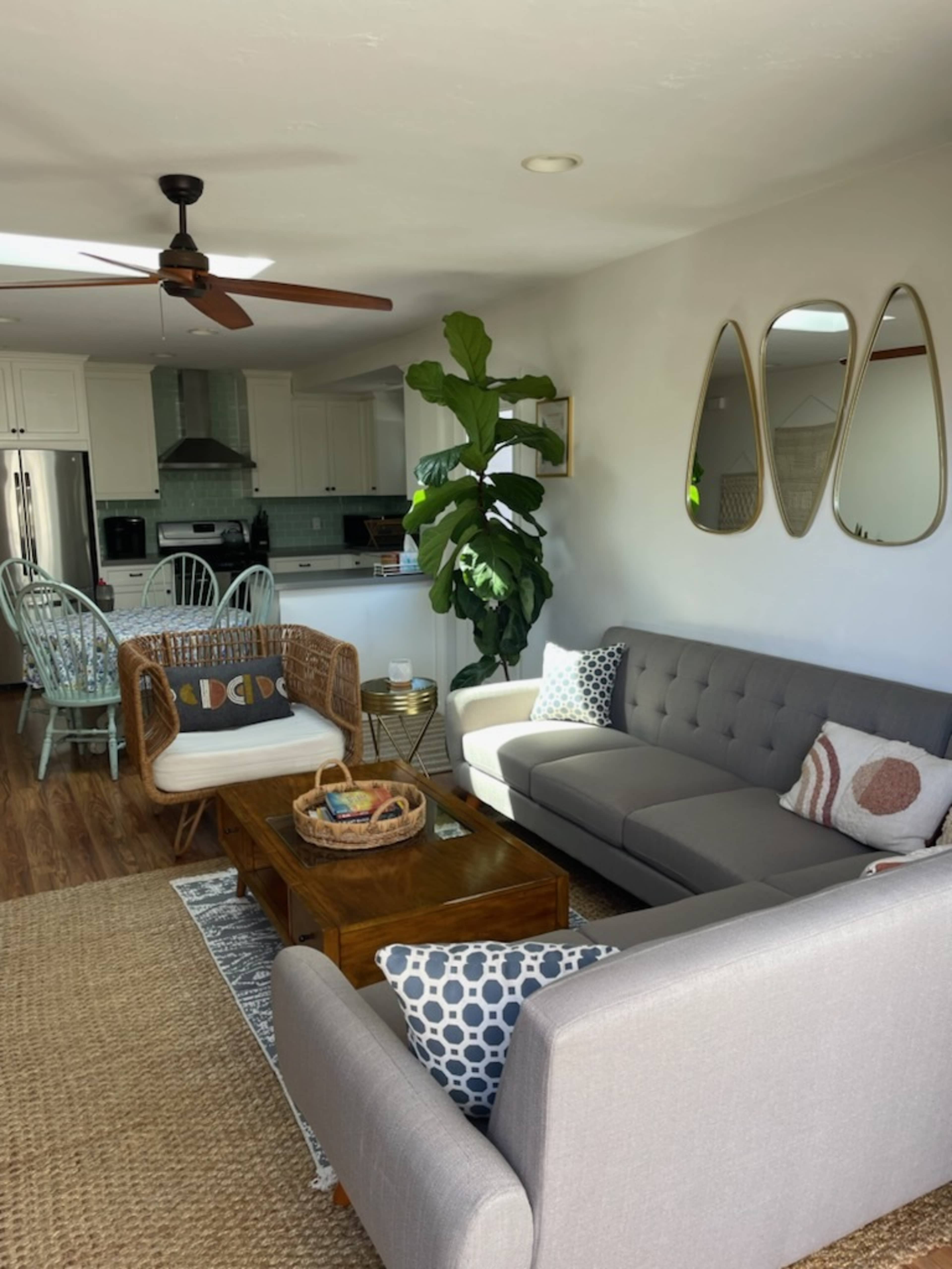 A bright living room featuring a gray sofa, a woven chair, a wooden coffee table, and a large plant, with a kitchen visible in the background.
