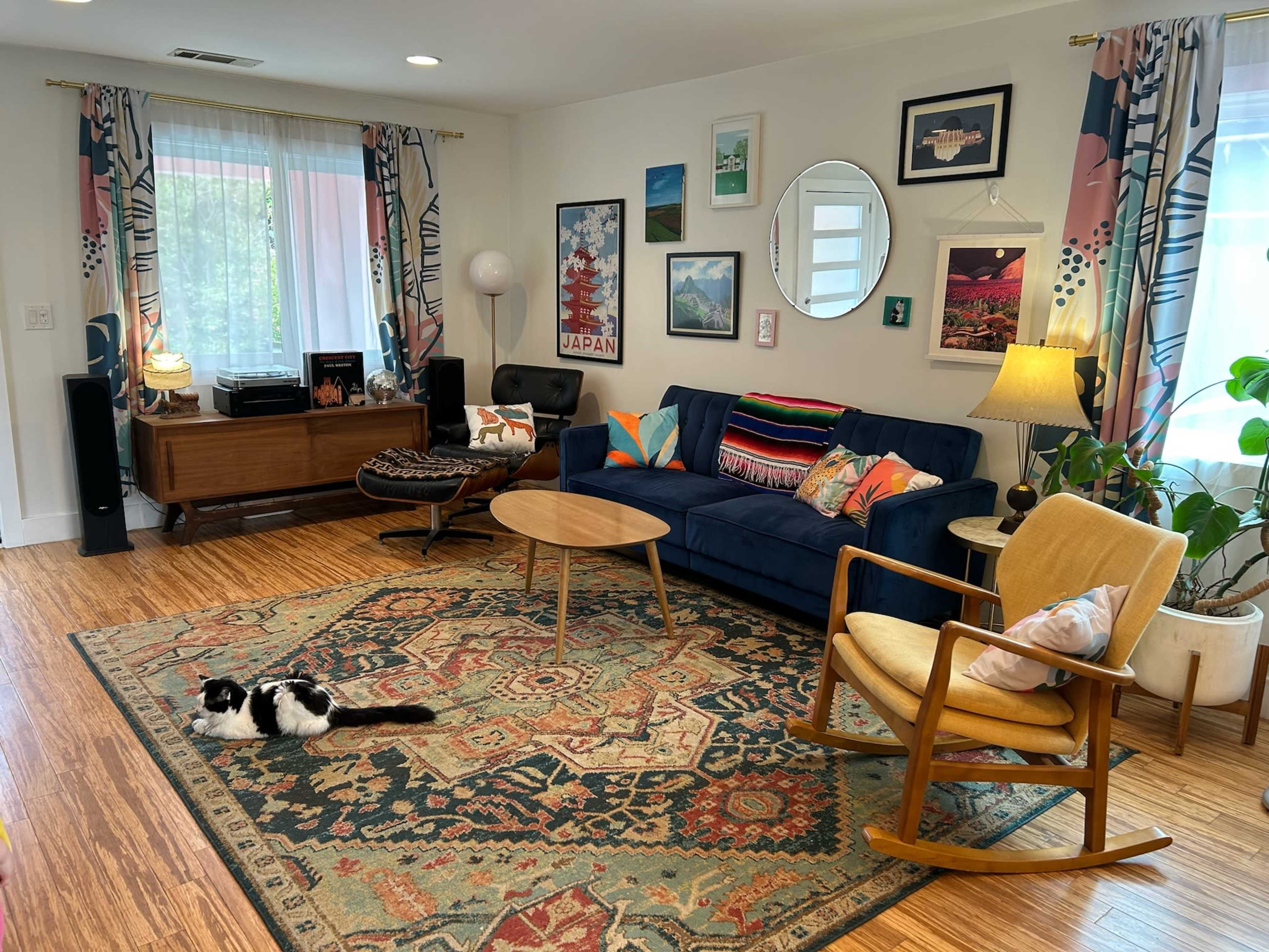 The room features a blue couch, a circular mirror, an assortment of artwork on the walls, and a cat resting on a patterned rug.