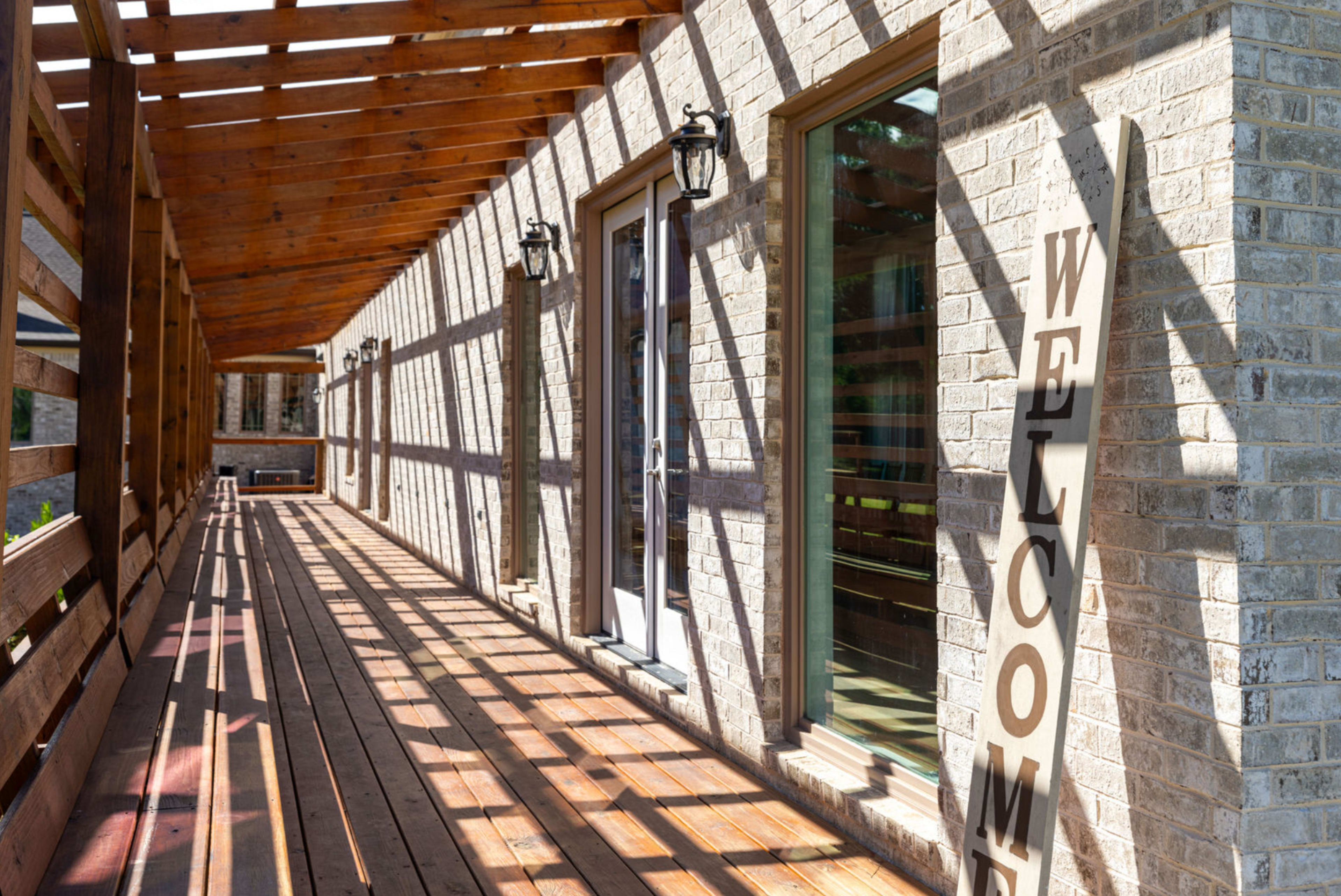The image shows a wooden veranda with a brick wall, large windows, and a decorative "WELCOME" sign leaning against the wall, with shadows created by the overhead beams.