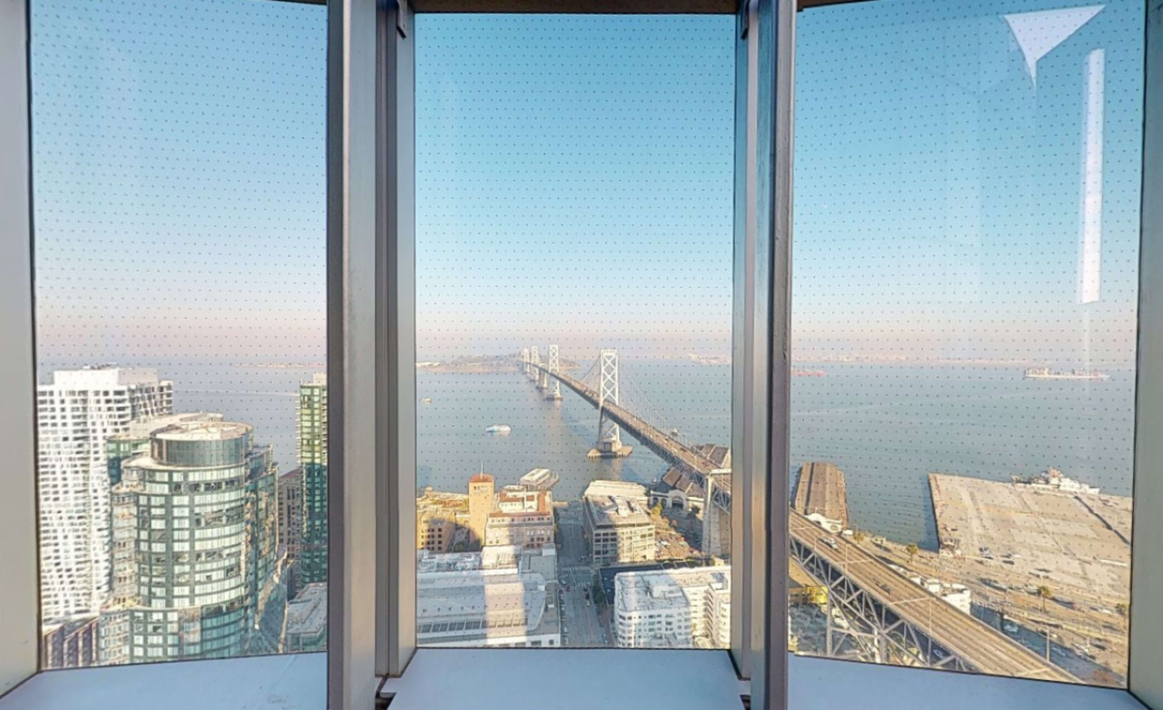 The image shows a panoramic view of the San Francisco Bay Bridge framed by glass panels from a high-rise building.