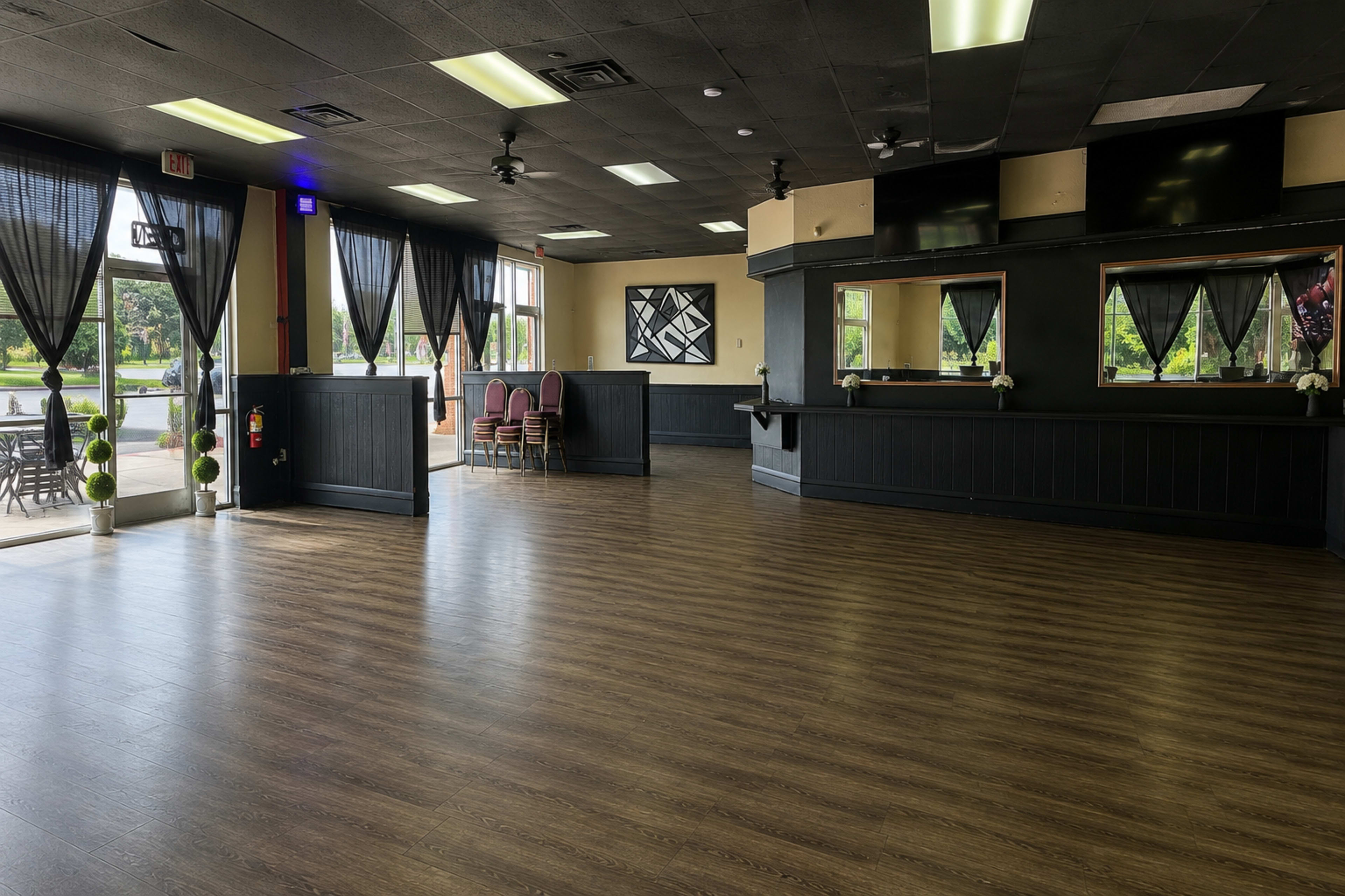 The interior of a spacious, empty event hall features dark walls, wooden flooring, and several chairs stacked in one corner under large windows with black curtains.