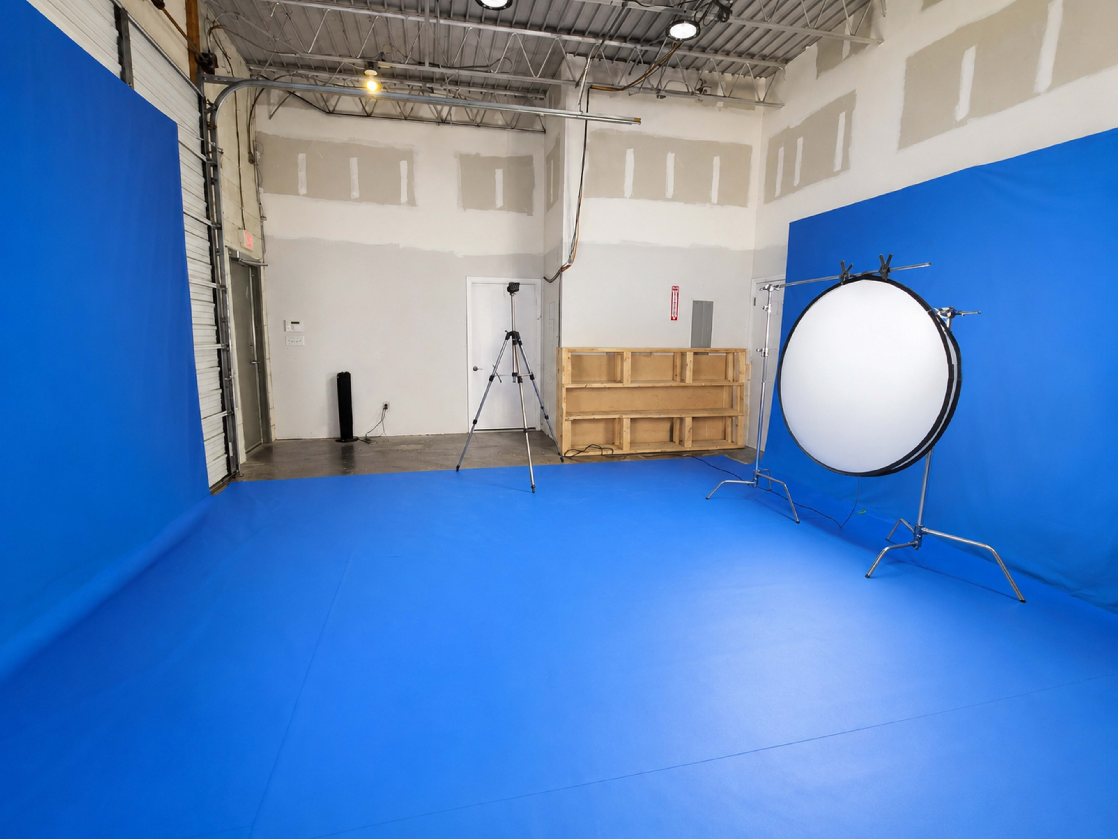 The image shows a studio space with a blue backdrop, a circular reflector on a stand, and a camera on a tripod set against a plain wall.