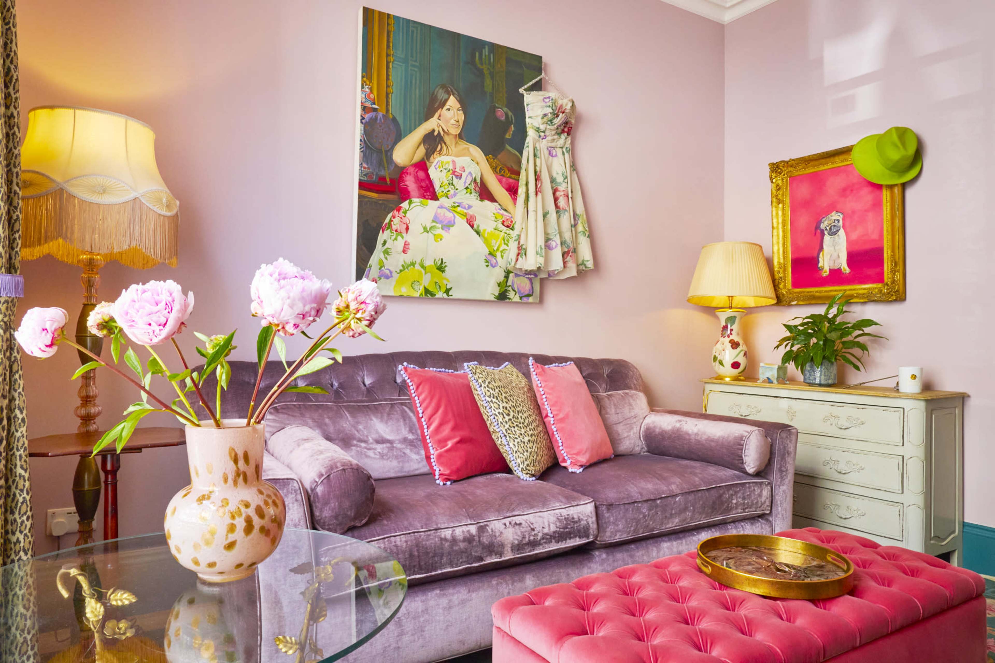A plush purple sofa with vibrant pillows is placed in a brightly colored room featuring floral artwork and a glass coffee table with a vase of pink peonies.