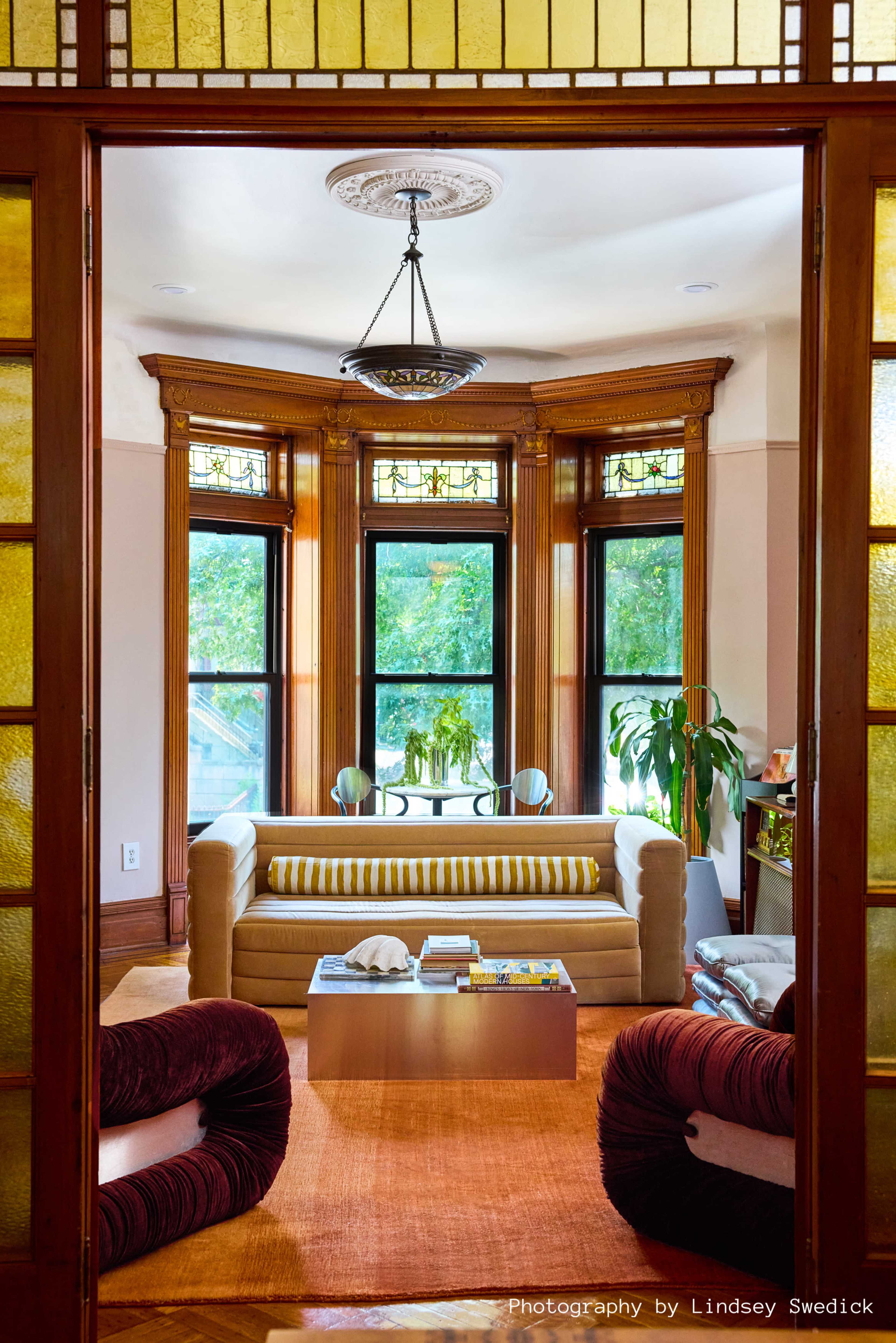 The image showcases a cozy living room with a bay window, featuring a beige sofa with yellow and white striped cushions, two red accent chairs, and a coffee table on an orange rug.