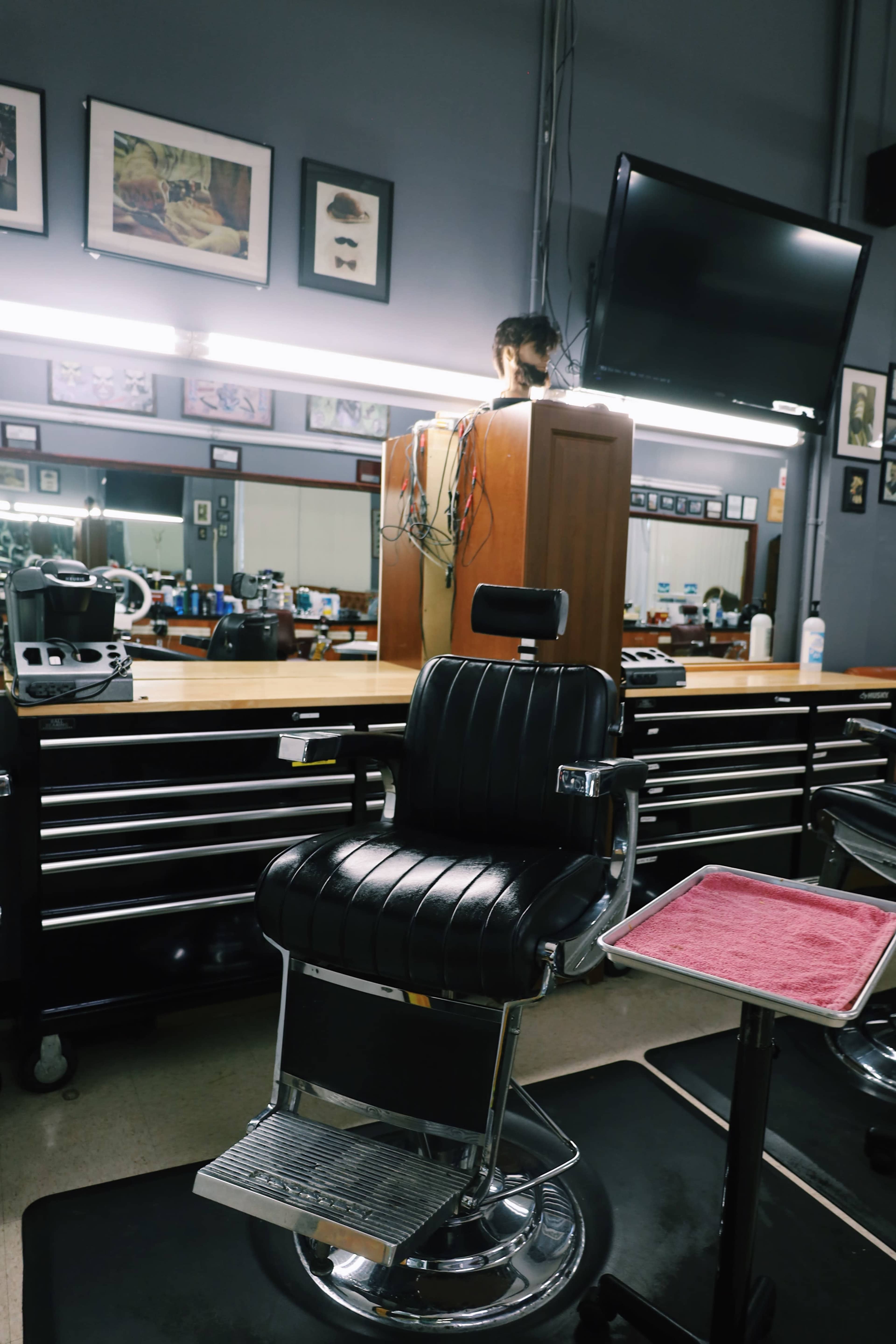 The image shows a barber shop interior with a black barber chair and a work station in the background, featuring a television and various tools.