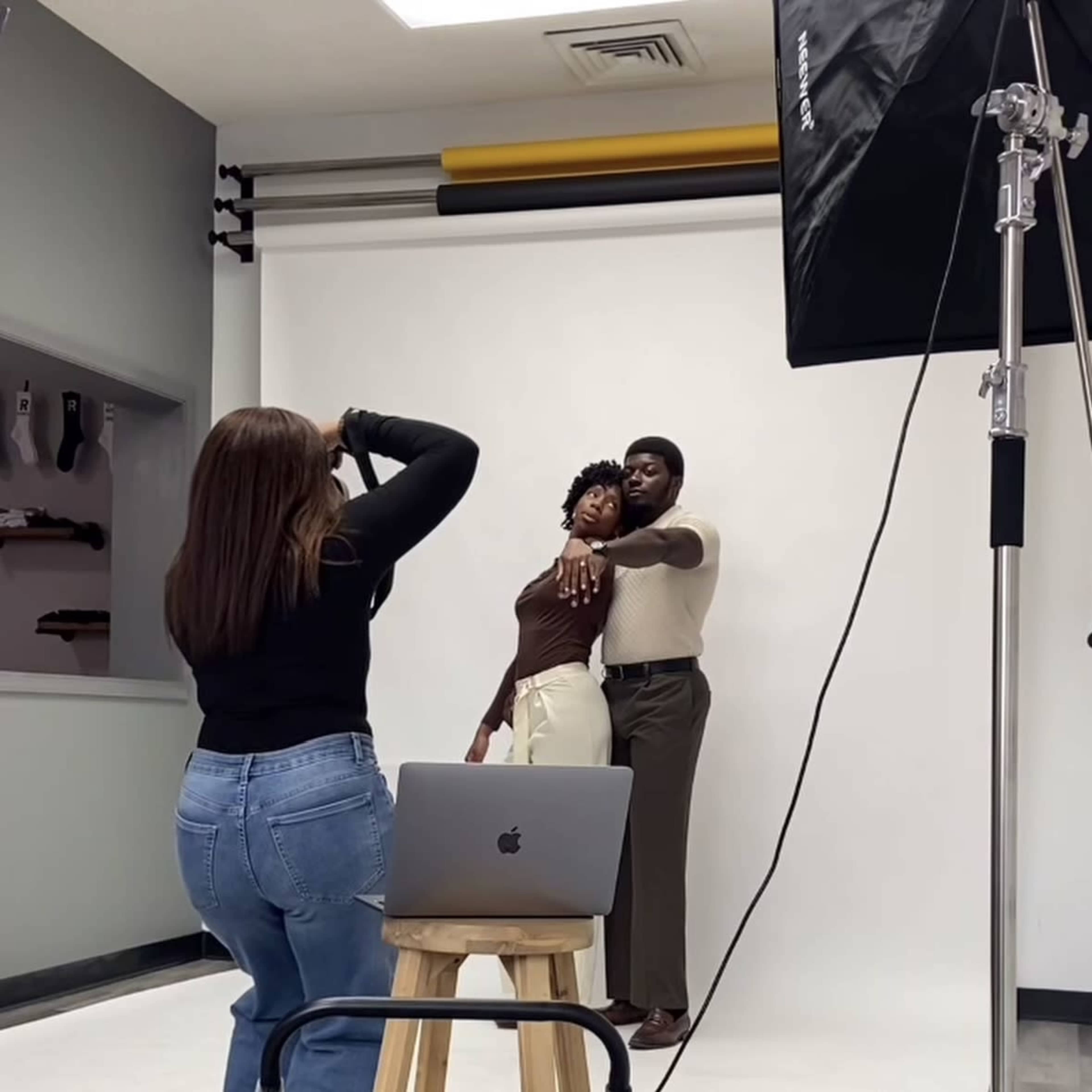 A photographer captures a couple posed in front of a plain white backdrop while using a laptop on a wooden stool.
