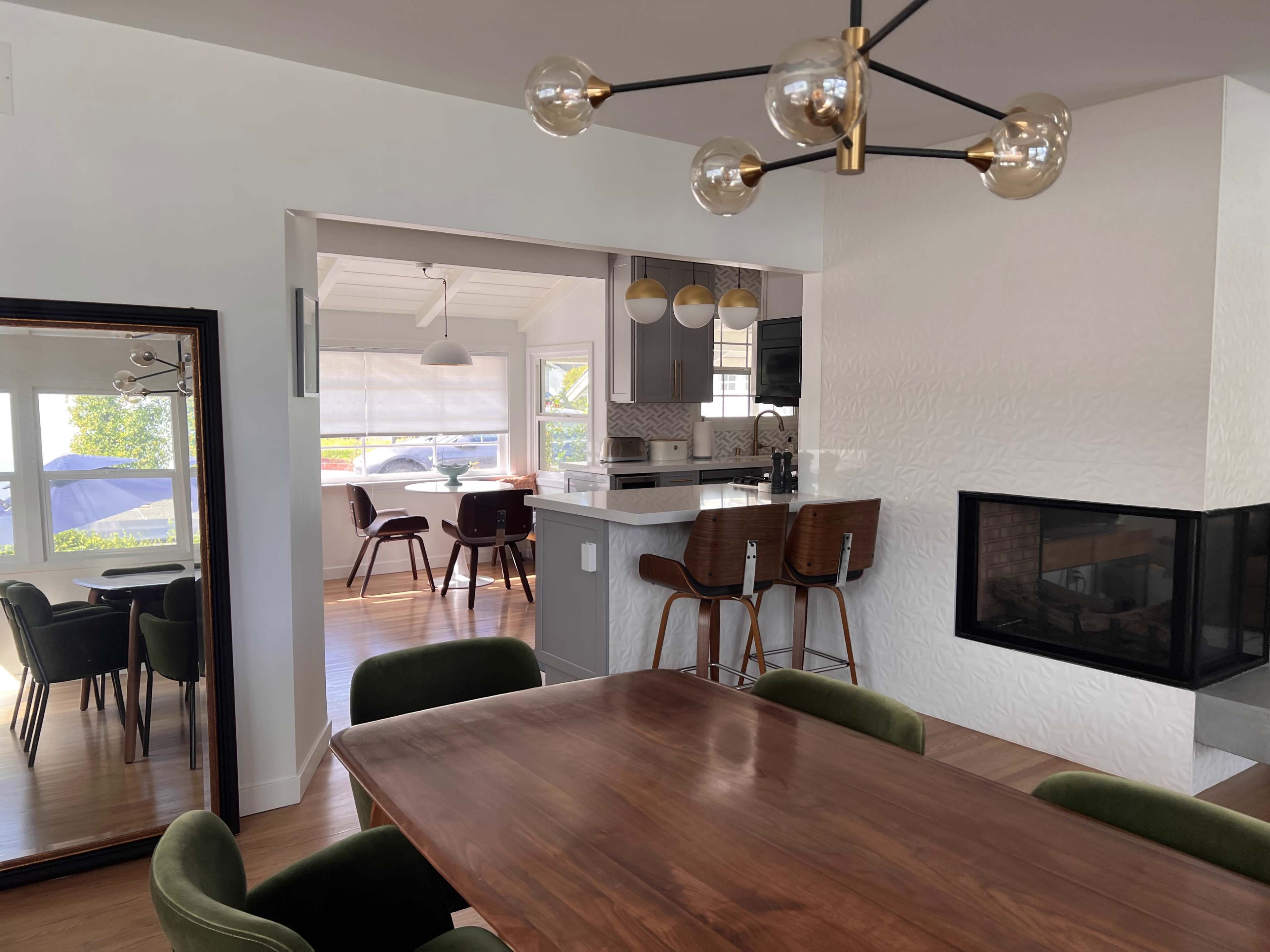 A modern open-concept living space featuring a dining area with a wooden table, green chairs, and a kitchen visible in the background with bar stools and a fireplace.