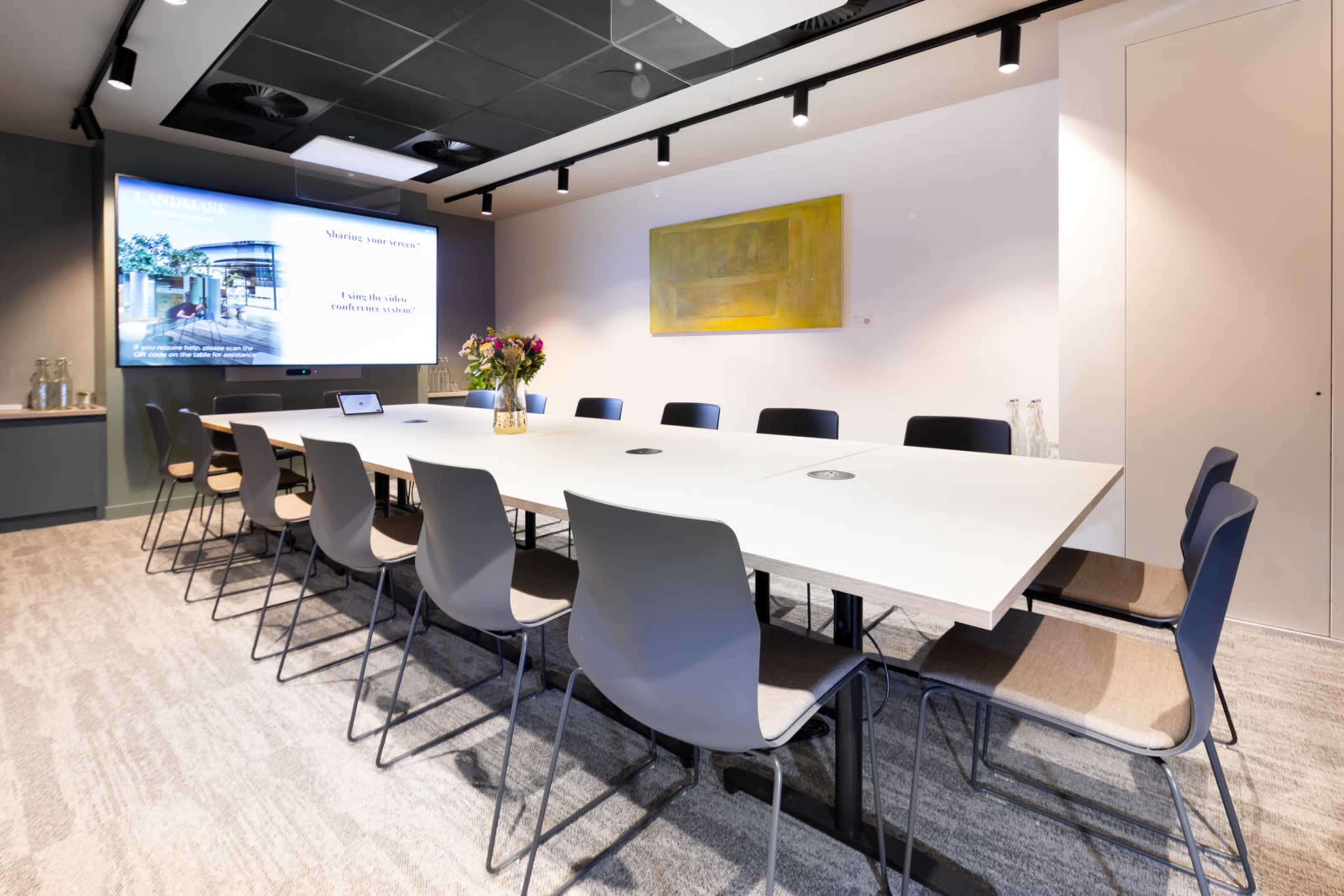 A modern conference room features a long table surrounded by chairs, with a large screen displaying a presentation and a vase of flowers on the table.